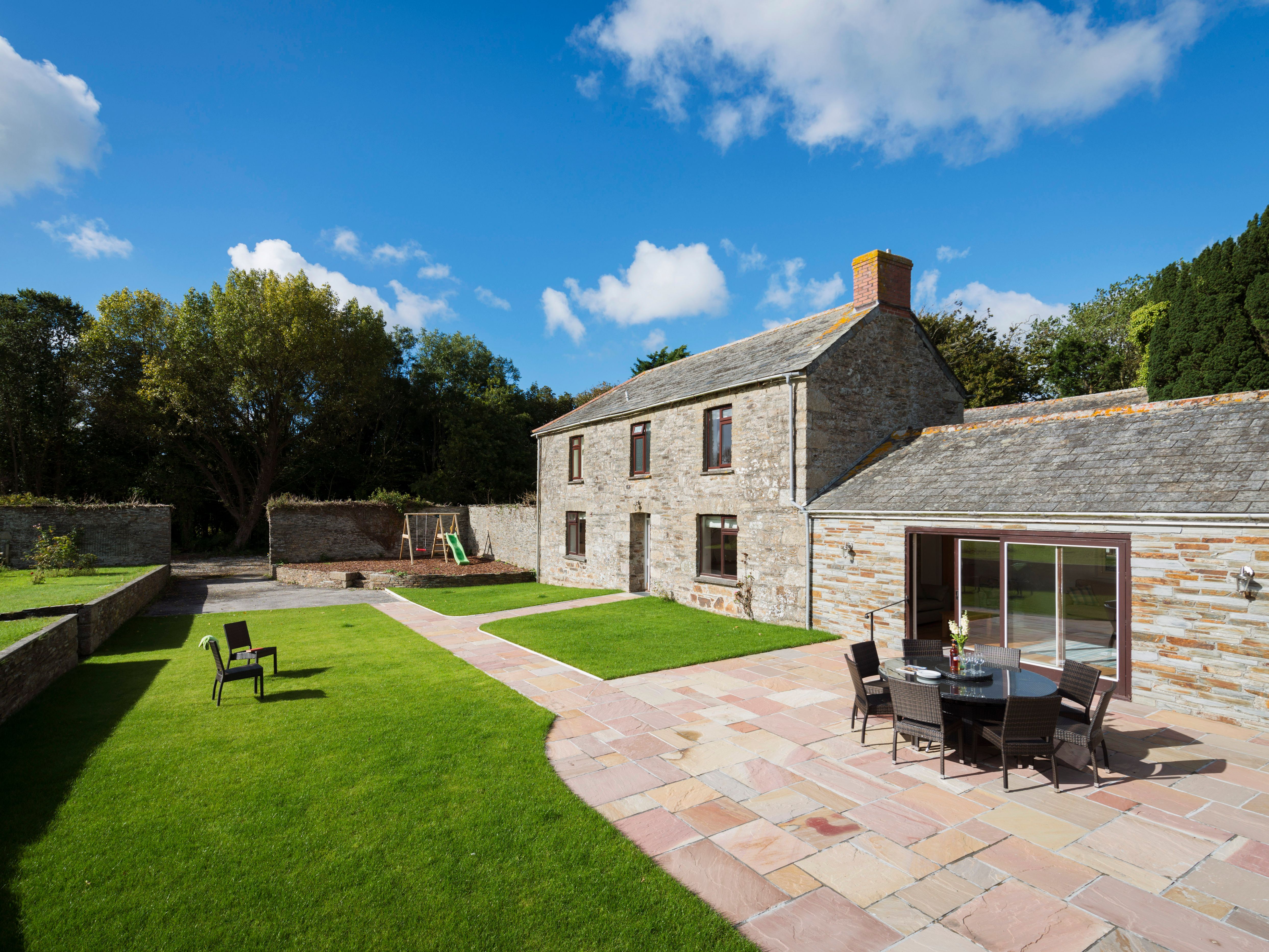 A traditional stone farmhouse with a well-maintained green lawn, paved patio, outdoor dining table and chairs, backed by trees and a blue sky with scattered clouds.