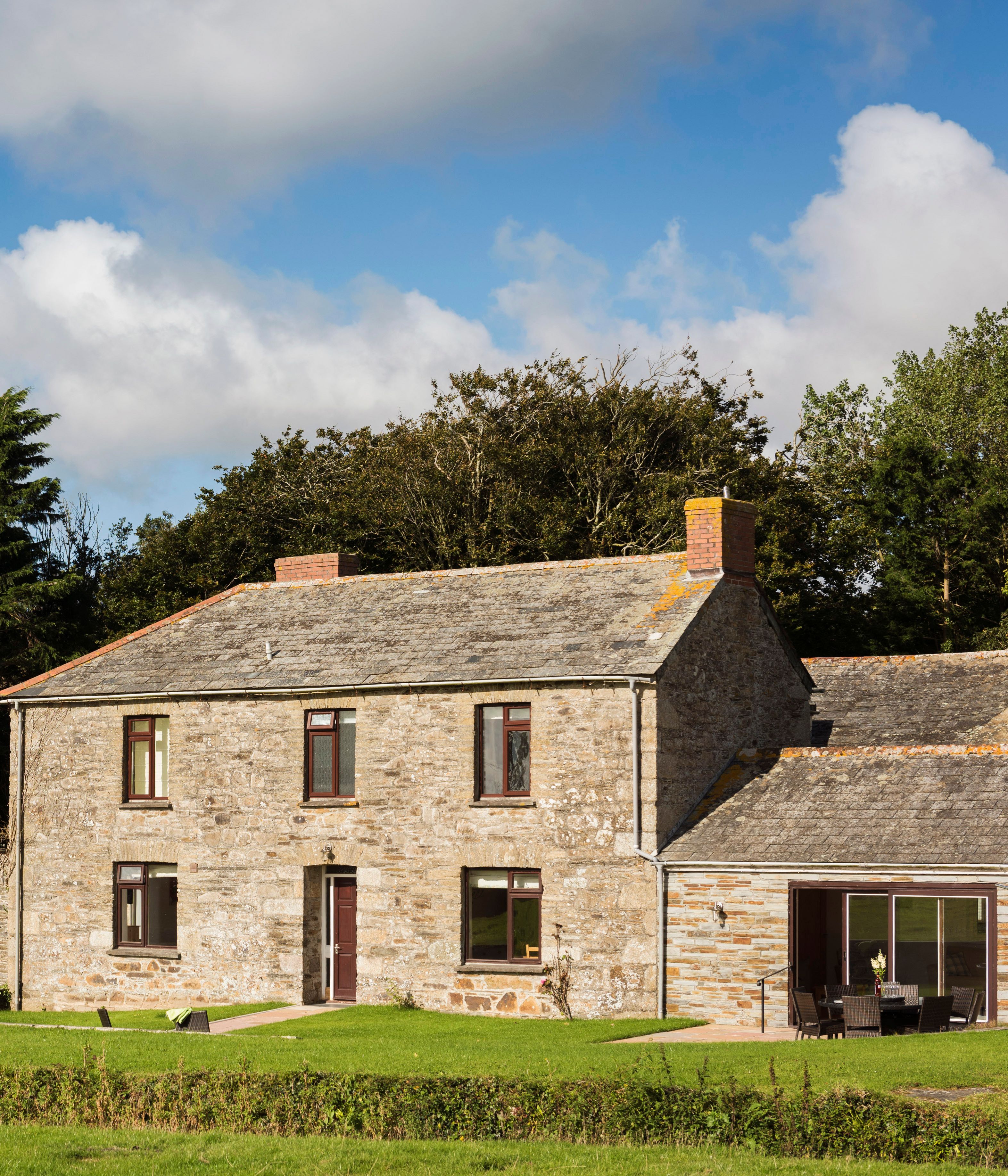 Traditional stone farmhouse with a grey slate roof, surrounded by green lawn and trees, with a children's playset and a stone wall in the garden under a blue sky with clouds.