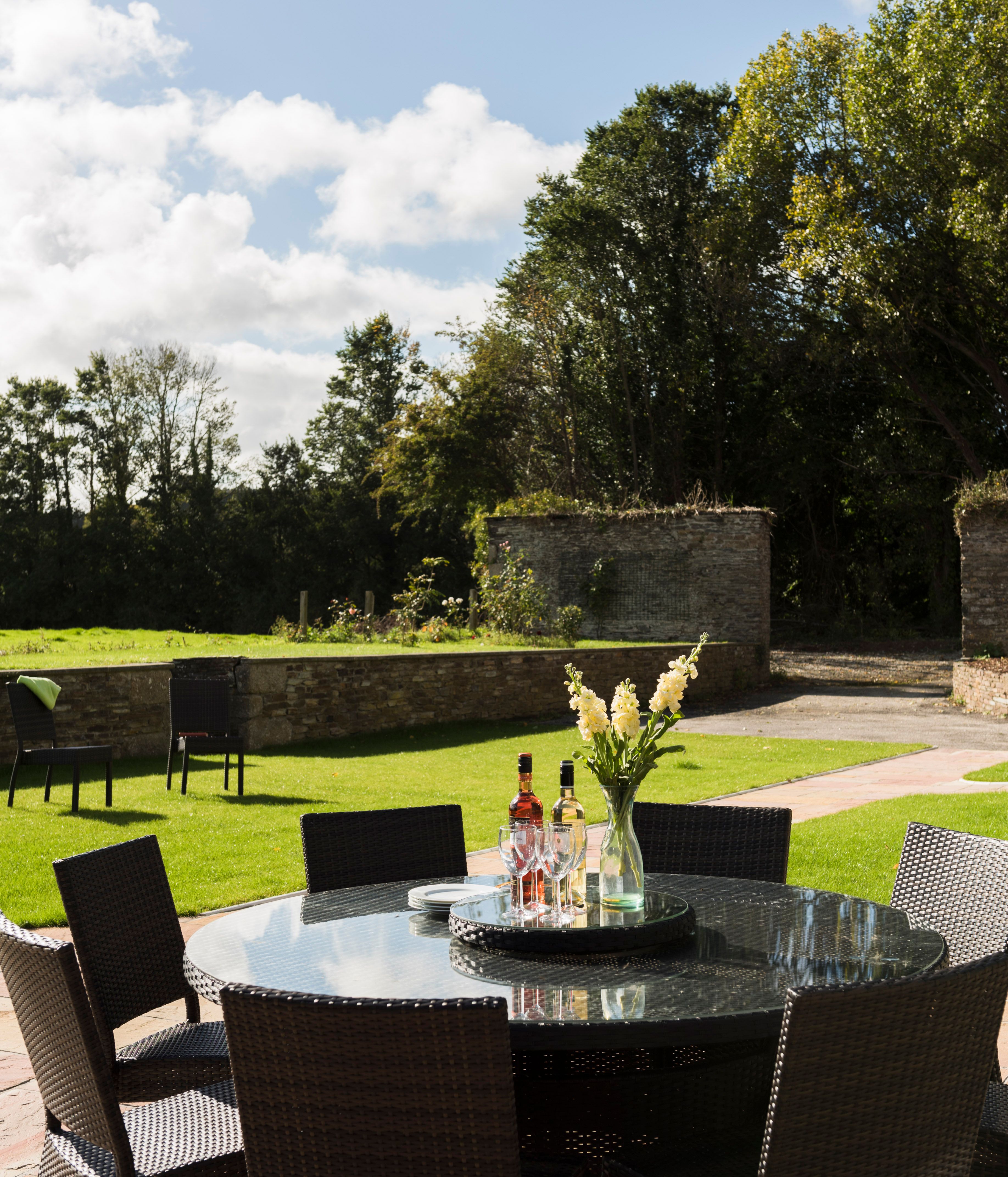 Outdoor patio with a round glass-top table surrounded by wicker chairs, set with bottles, glasses, and a vase of yellow flowers, overlooking a grassy garden with trees and a stone wall.