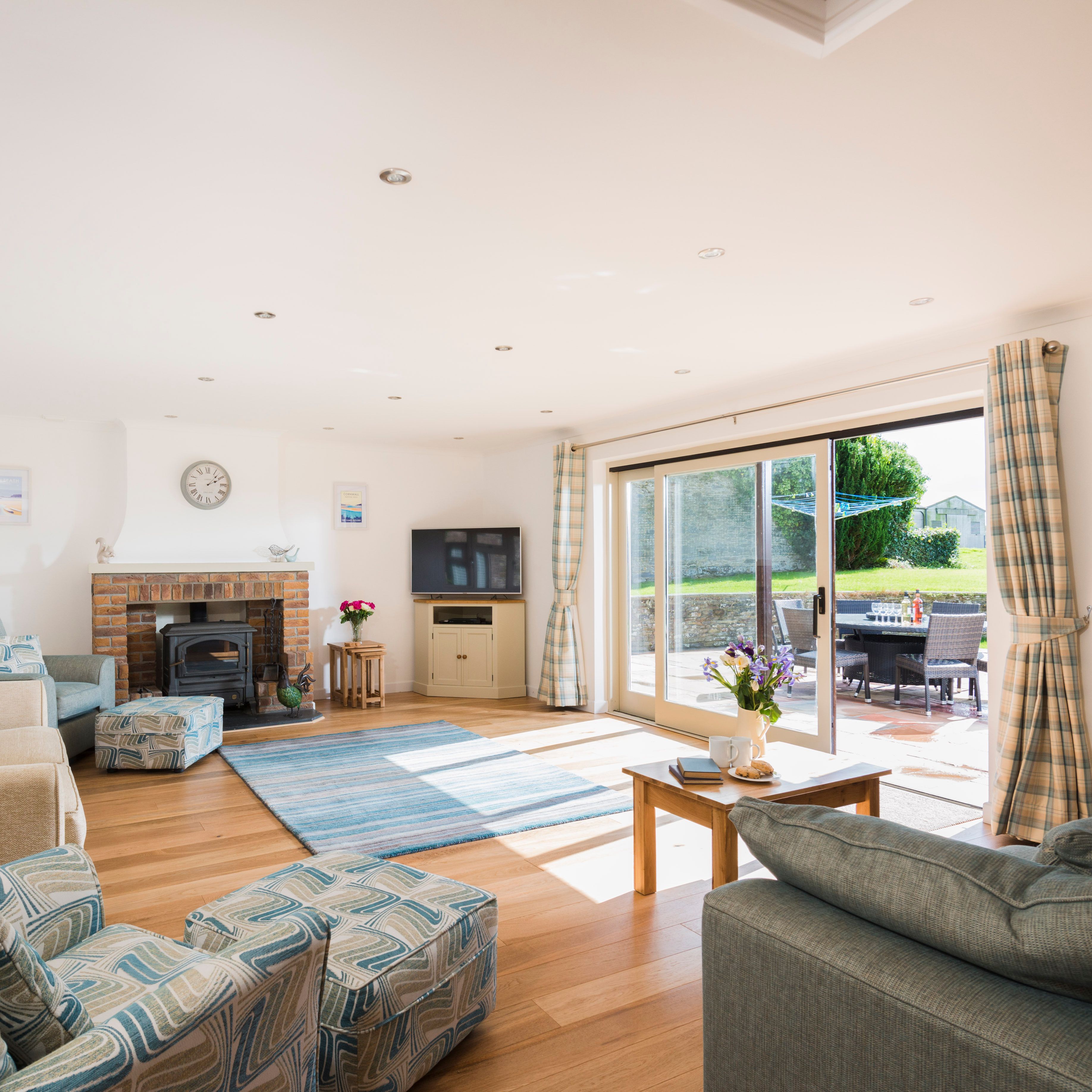 Bright and spacious living room with large windows, cozy sofas, a brick fireplace, television, patterned armchairs, and sliding glass doors opening to a sunny patio with outdoor seating.