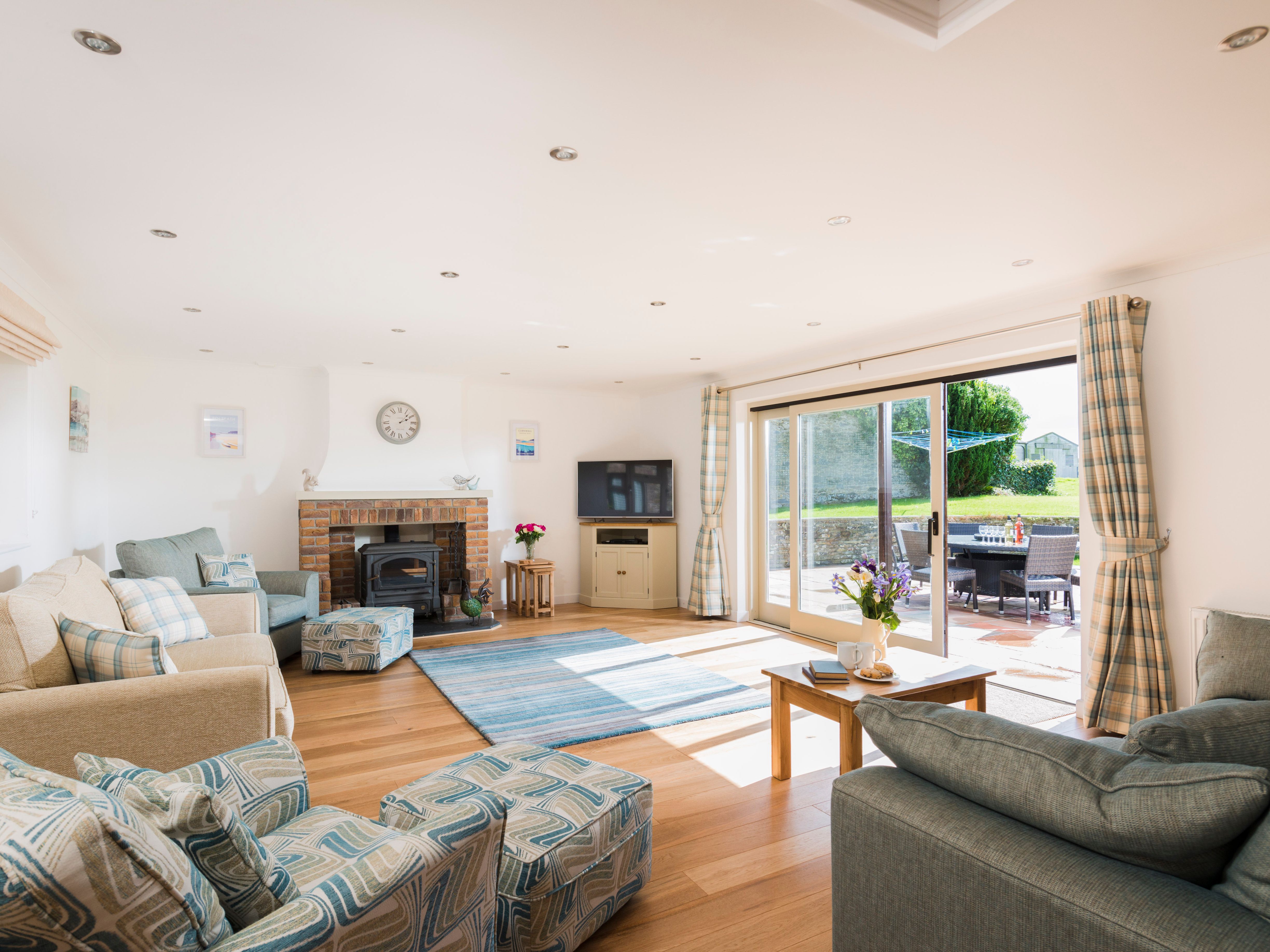 Bright and spacious living room with large windows, cozy sofas, a brick fireplace, television, patterned armchairs, and sliding glass doors opening to a sunny patio with outdoor seating.