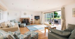 Bright and spacious living room with large windows, cozy sofas, a brick fireplace, television, patterned armchairs, and sliding glass doors opening to a sunny patio with outdoor seating.