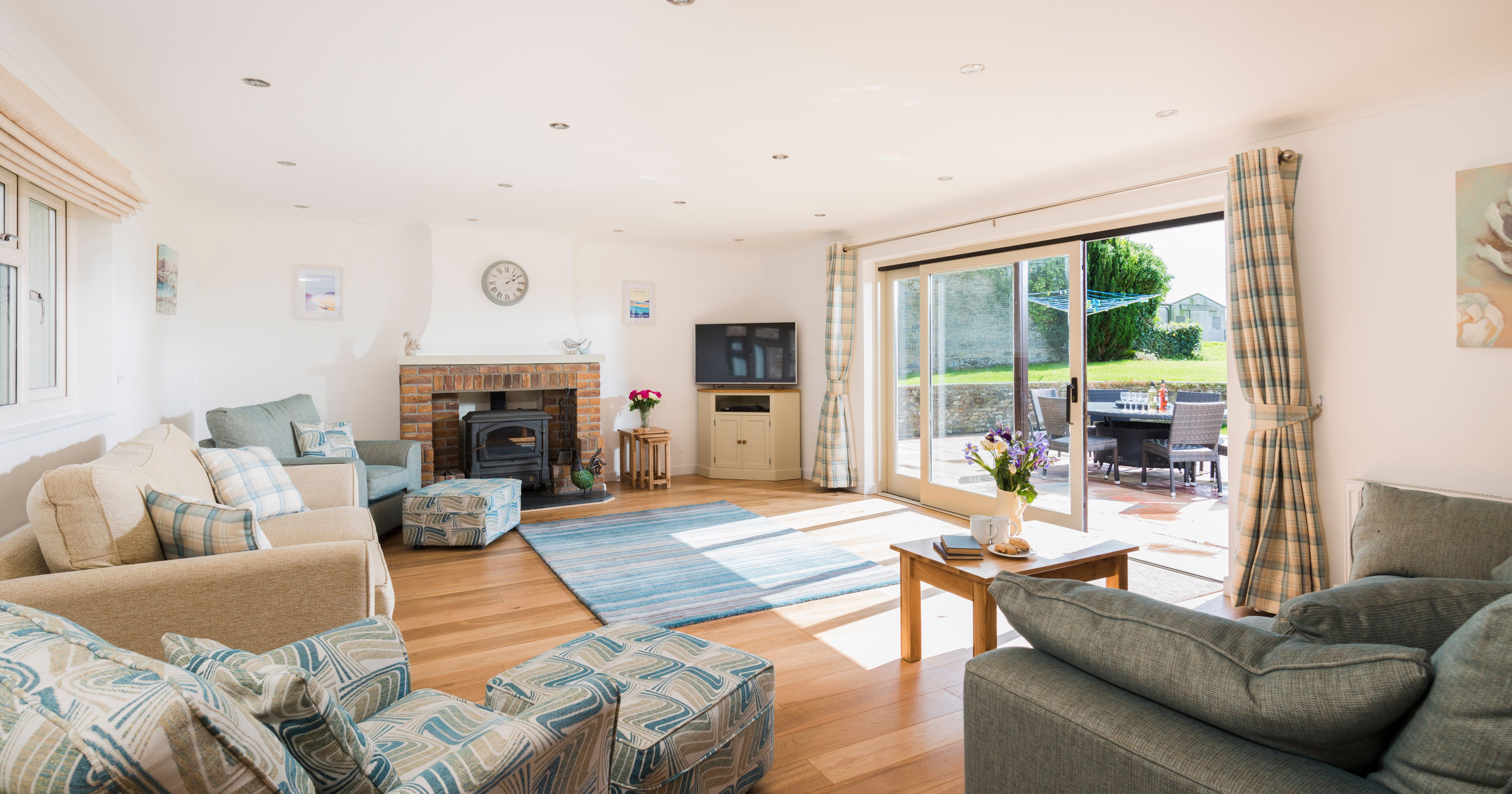 Bright and spacious living room with large windows, cozy sofas, a brick fireplace, television, patterned armchairs, and sliding glass doors opening to a sunny patio with outdoor seating.