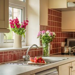 Bright kitchen with red tile backsplash, stainless sink, fresh flowers in vases, and a chopping board with strawberries and pomegranate on the countertop.