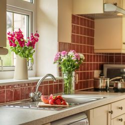 Bright kitchen with red tile backsplash, stainless sink, fresh flowers in vases, and a chopping board with strawberries and pomegranate on the countertop.