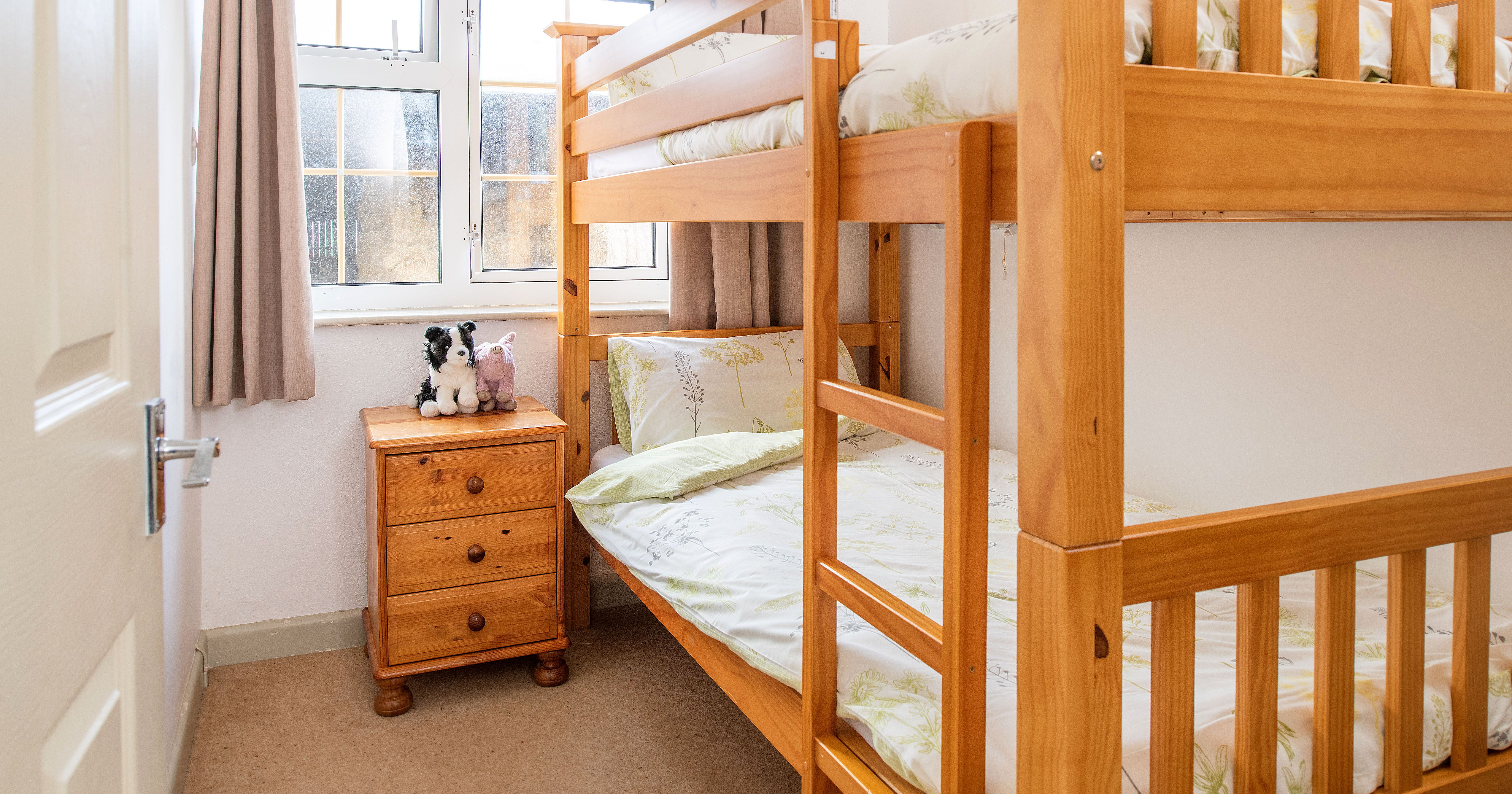 Small bedroom with wooden bunk beds, a wooden nightstand with drawers, two stuffed animals on the nightstand, and a window with beige curtains.