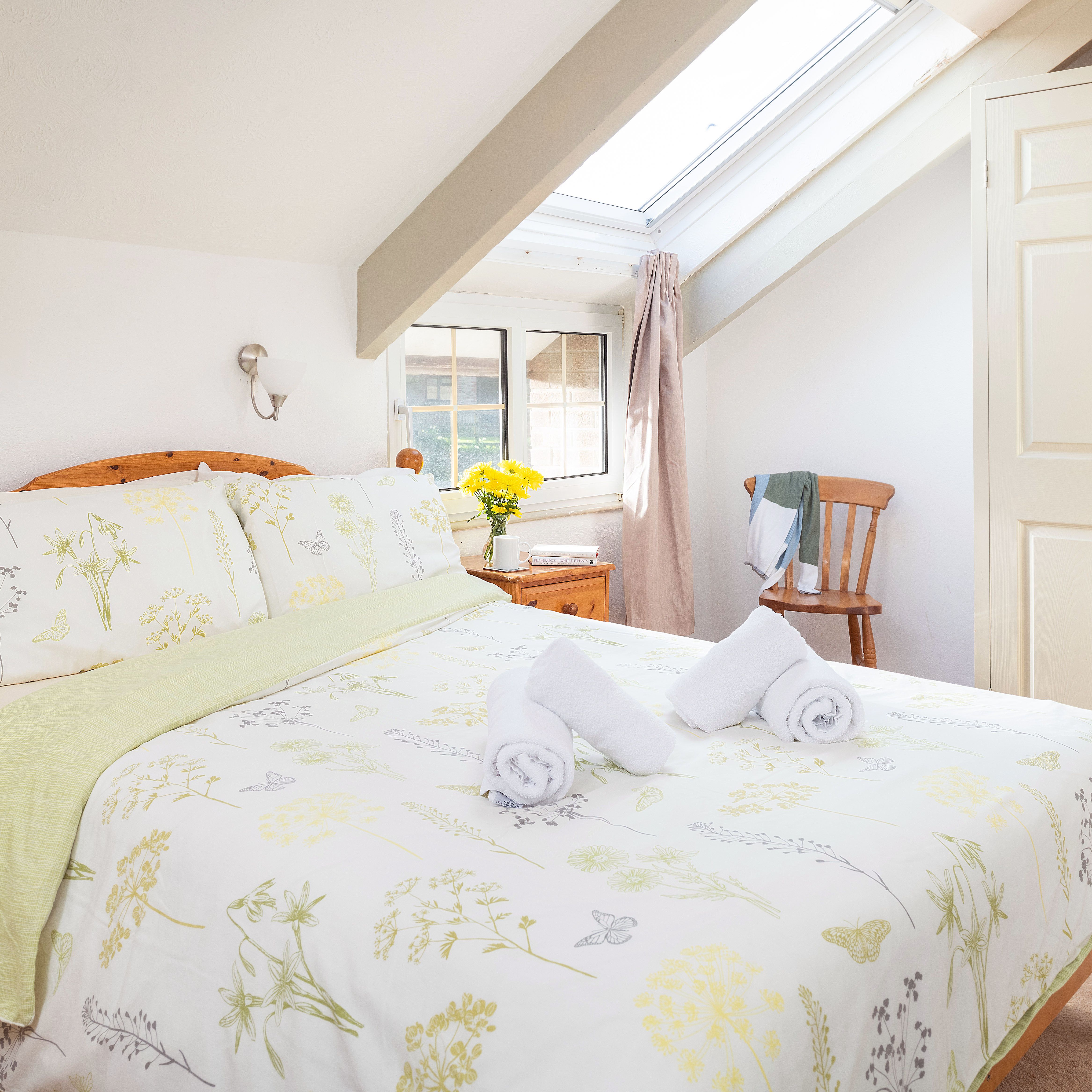 Bright attic bedroom with wooden furniture, floral bedding, two rolled towels on the bed, skylight window, and a small nightstand with a cup and a vase of yellow flowers.
