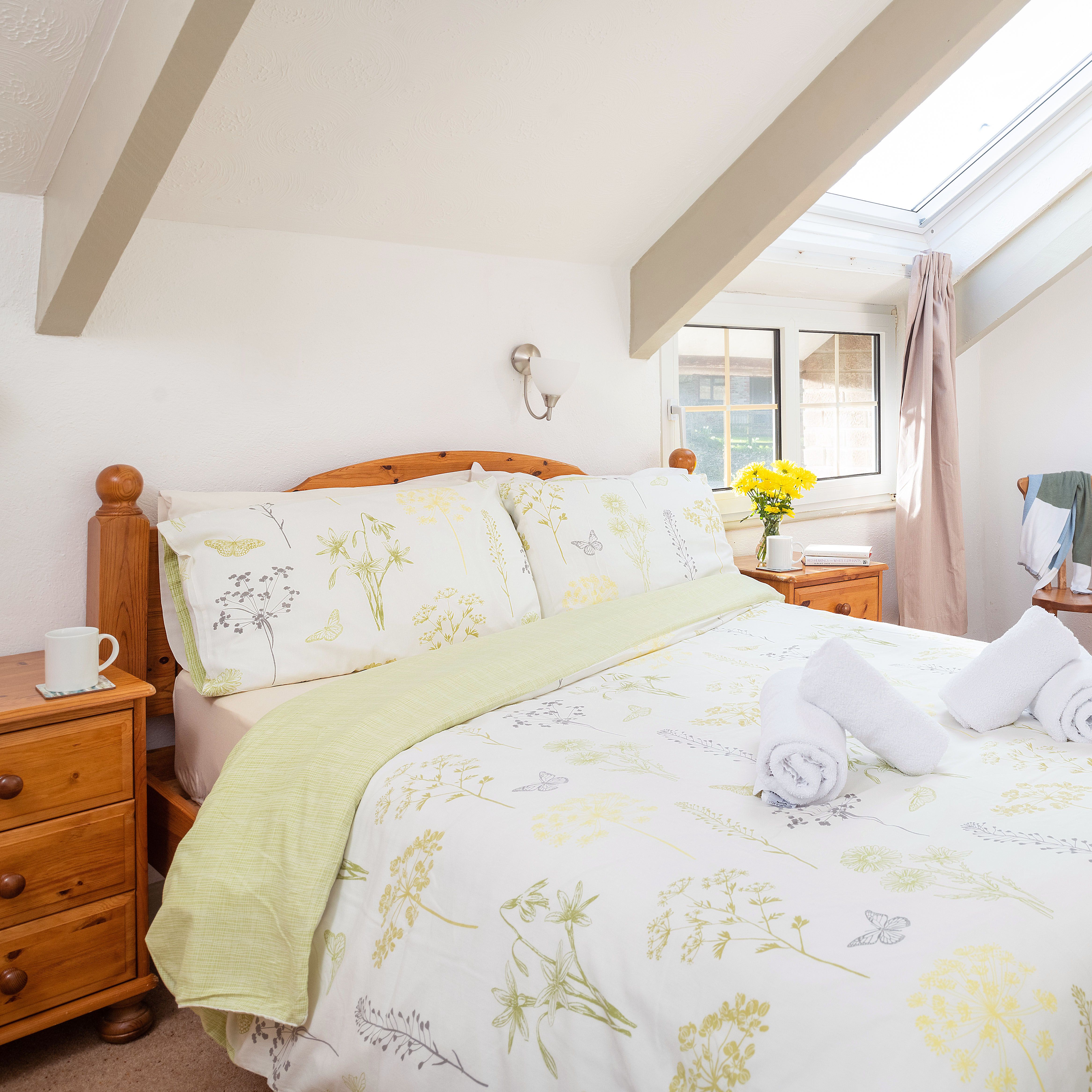 Bright attic bedroom with wooden furniture, floral bedding, two rolled towels on the bed, skylight window, and a small nightstand with a cup and a vase of yellow flowers.