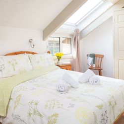 Bright attic bedroom with wooden furniture, floral bedding, two rolled towels on the bed, skylight window, and a small nightstand with a cup and a vase of yellow flowers.