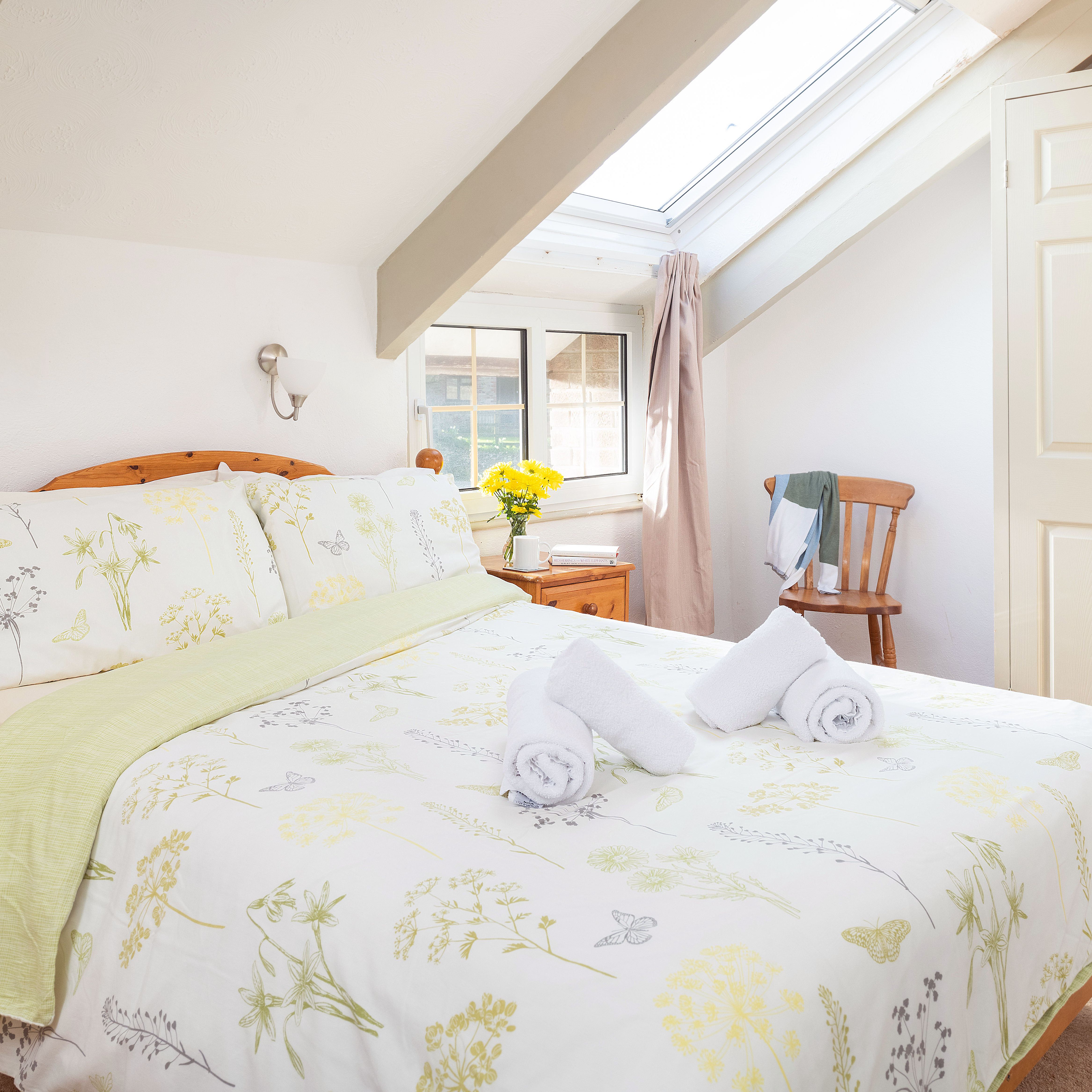 Bright attic bedroom with wooden furniture, floral bedding, two rolled towels on the bed, skylight window, and a small nightstand with a cup and a vase of yellow flowers.