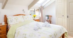 Bright attic bedroom with wooden furniture, floral bedding, two rolled towels on the bed, skylight window, and a small nightstand with a cup and a vase of yellow flowers.