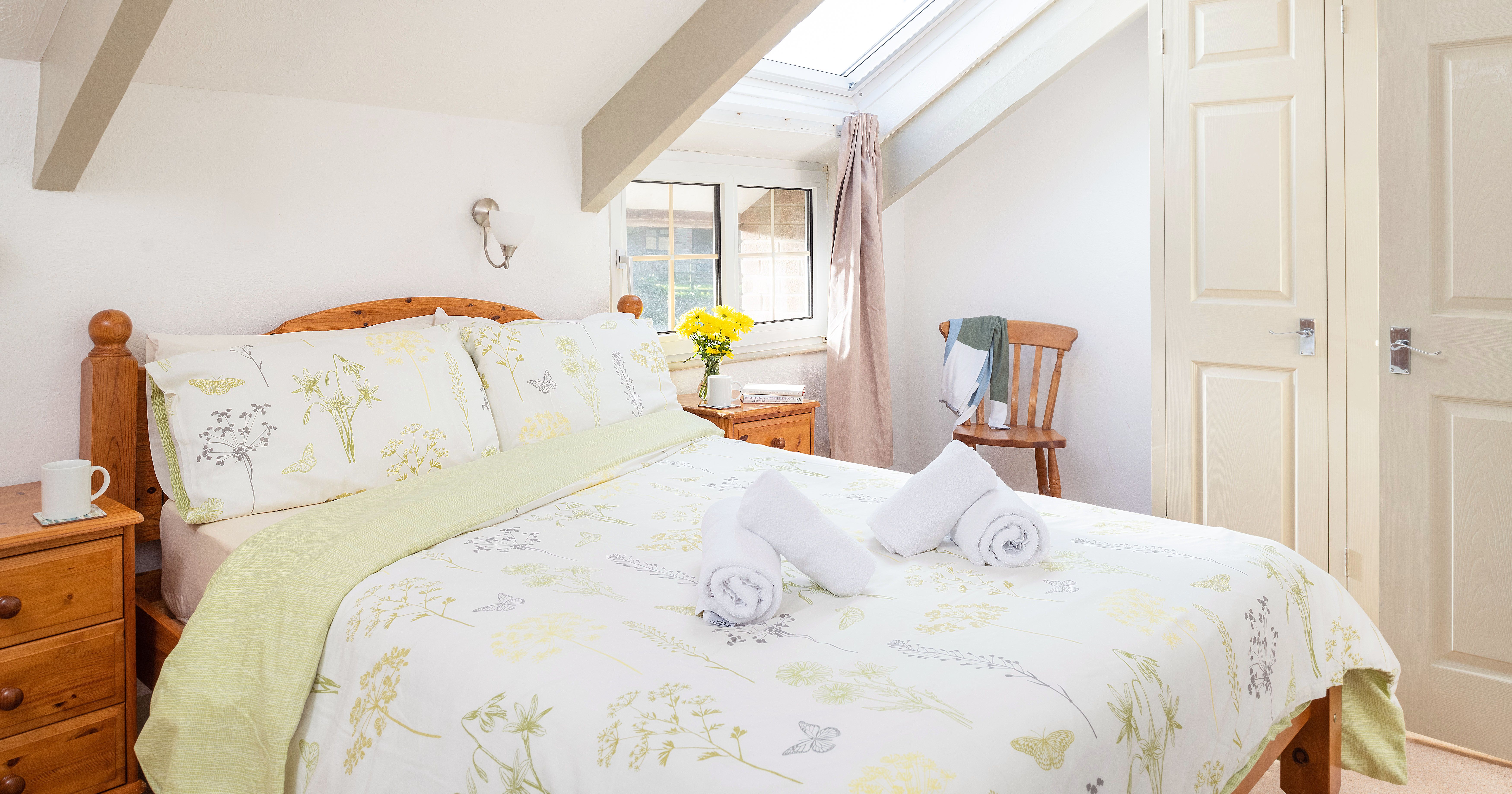 Bright attic bedroom with wooden furniture, floral bedding, two rolled towels on the bed, skylight window, and a small nightstand with a cup and a vase of yellow flowers.