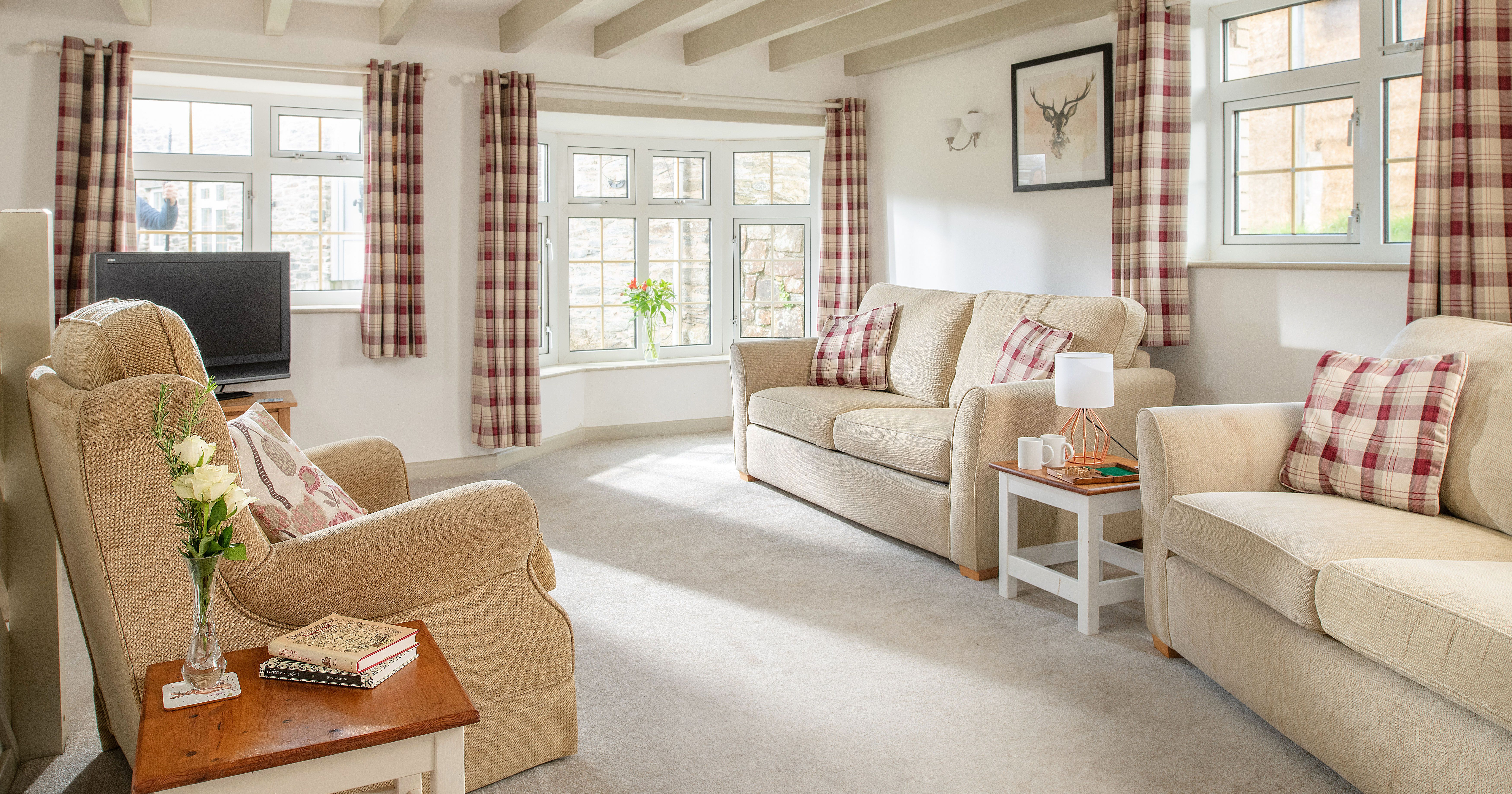 Bright and cozy living room with beige sofas, plaid curtains, a TV, and a side table with flowers and books. Large windows let in natural light.