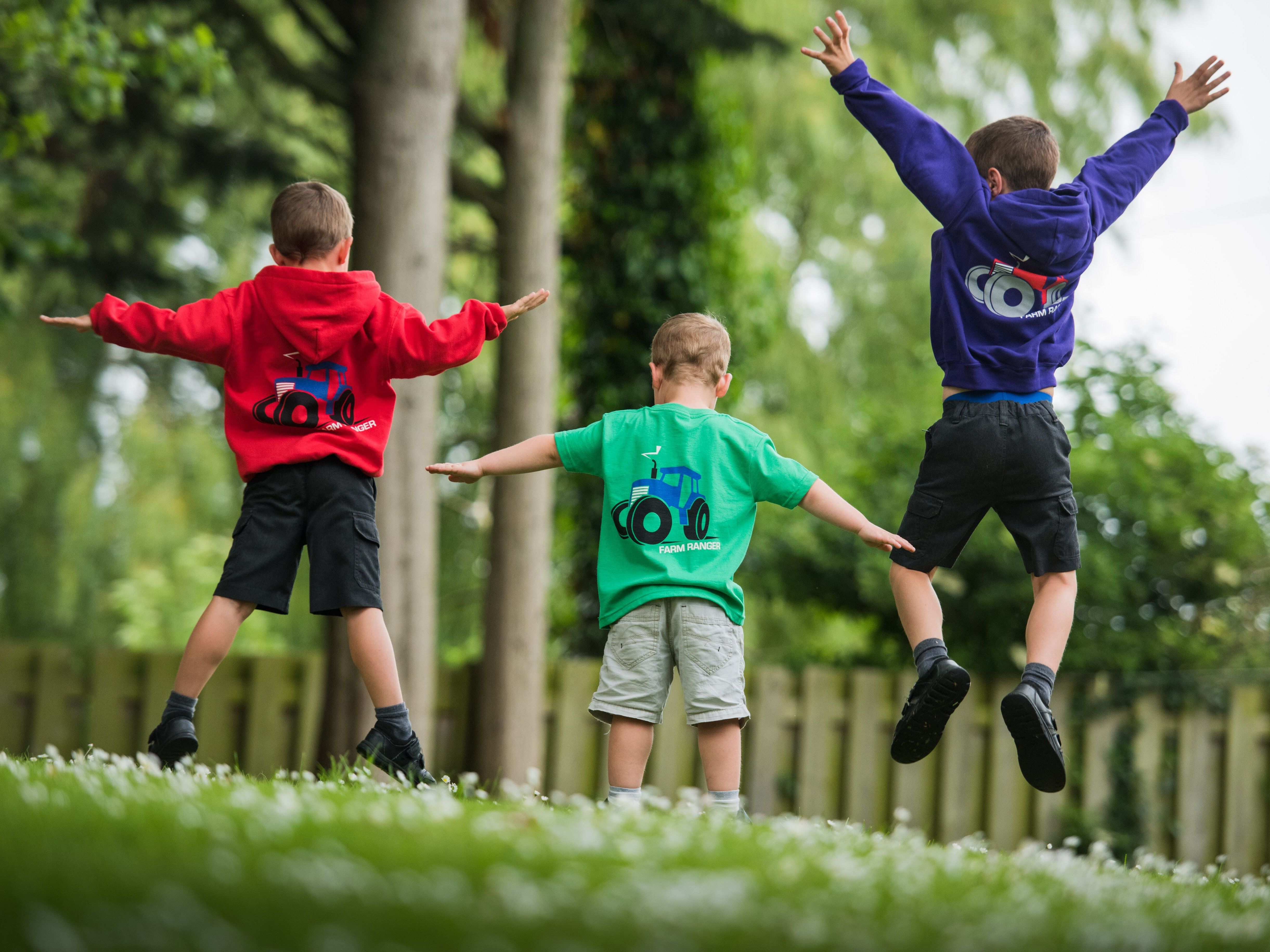 Three children seen from behind, wearing colorful tops with tractor designs, playing and jumping outdoors on a grassy area near trees and a wooden fence.