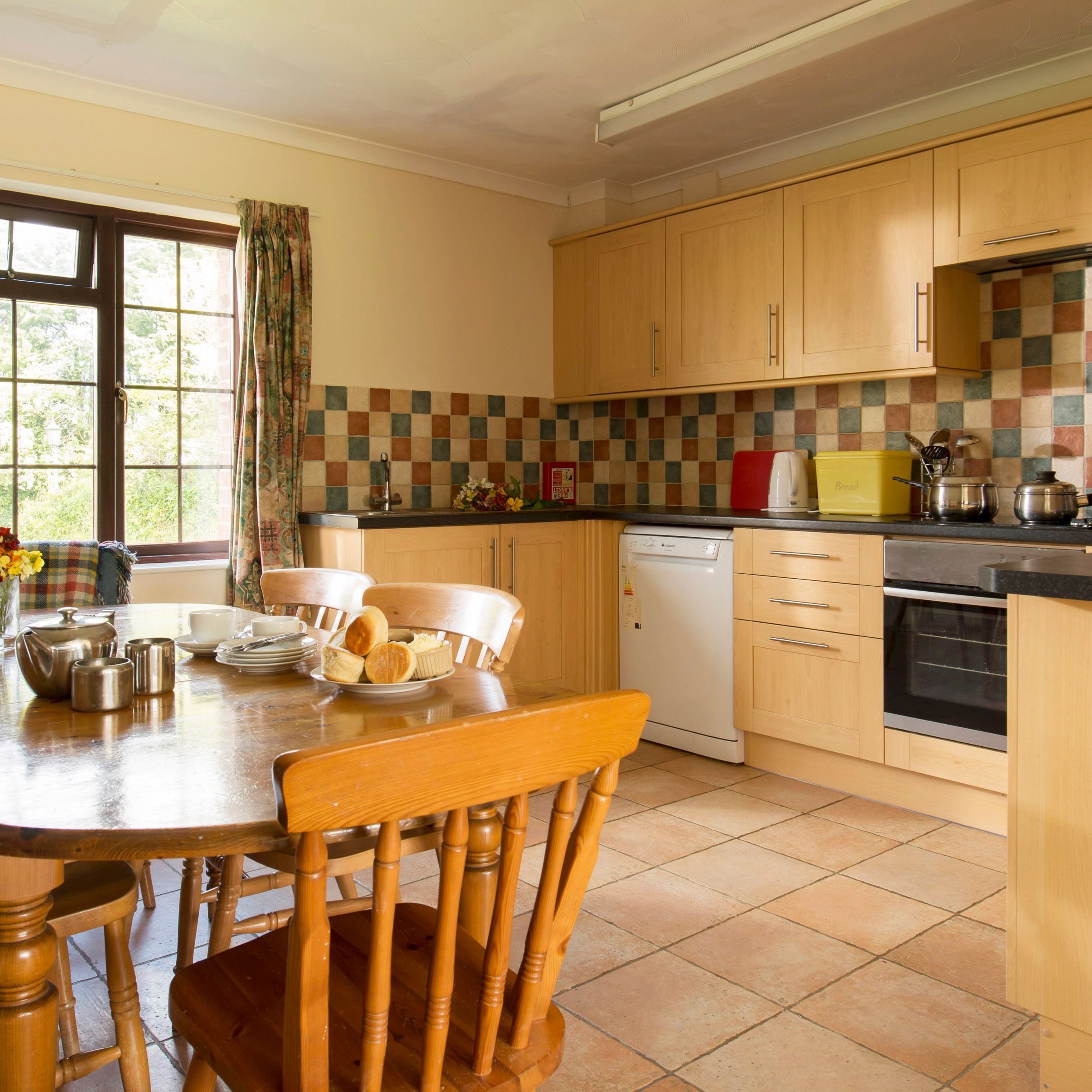 Cozy kitchen with wooden cabinets, a tiled backsplash, and a dining table set for tea with plates of pastries and a teapot. Large window provides natural light and view of greenery outside.