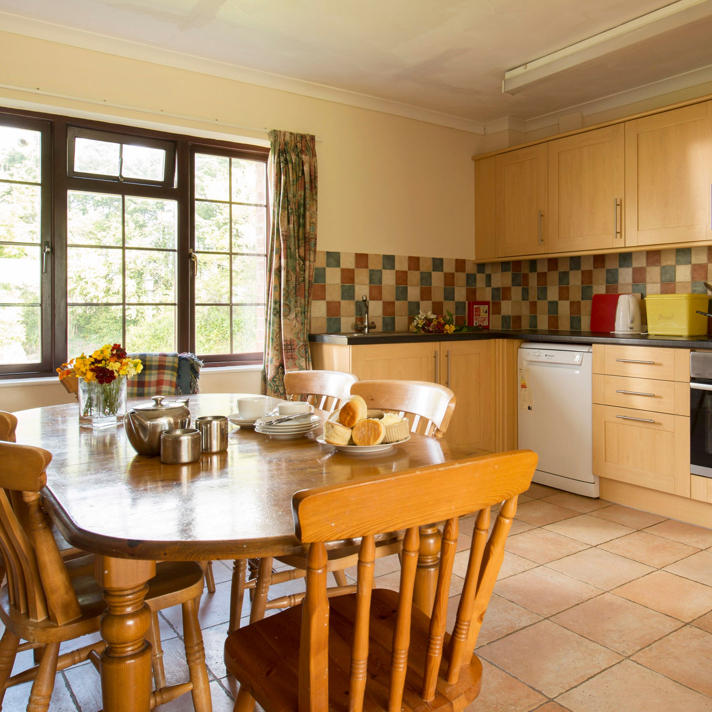 Cozy kitchen with wooden cabinets, a tiled backsplash, and a dining table set for tea with plates of pastries and a teapot. Large window provides natural light and view of greenery outside.