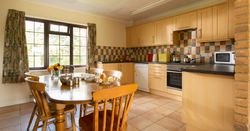 Cozy kitchen with wooden cabinets, a tiled backsplash, and a dining table set for tea with plates of pastries and a teapot. Large window provides natural light and view of greenery outside.