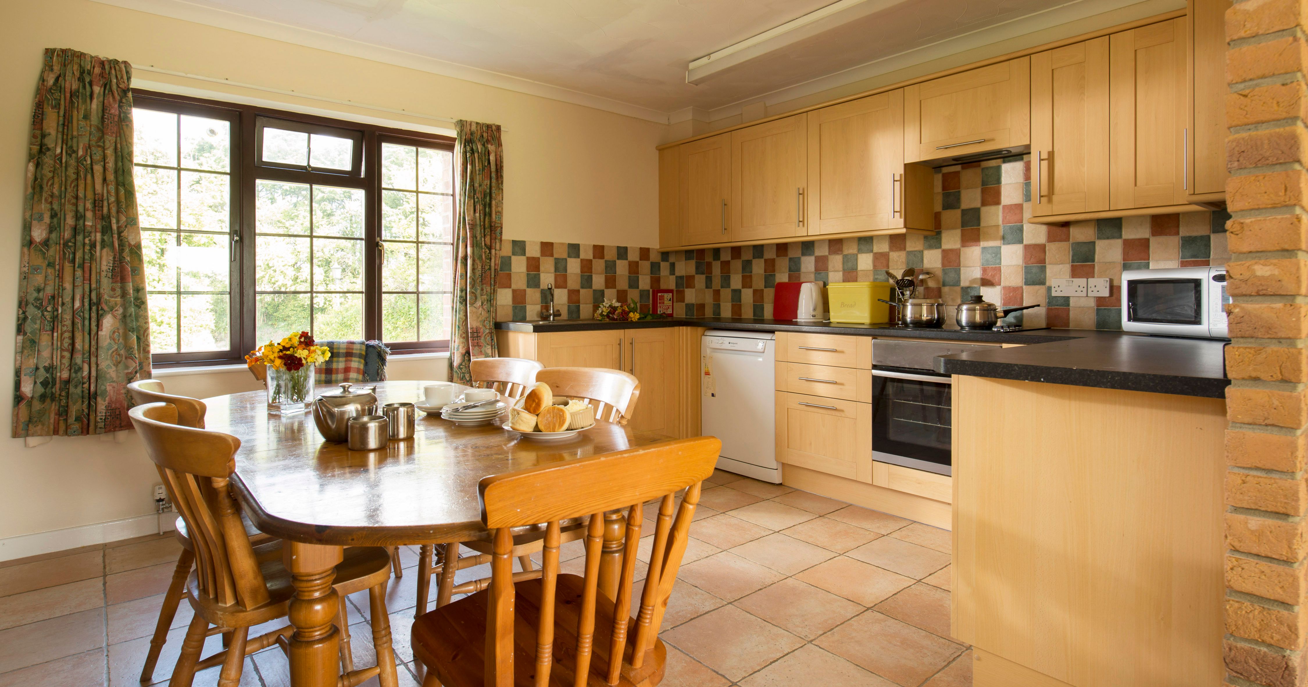 Cozy kitchen with wooden cabinets, a tiled backsplash, and a dining table set for tea with plates of pastries and a teapot. Large window provides natural light and view of greenery outside.