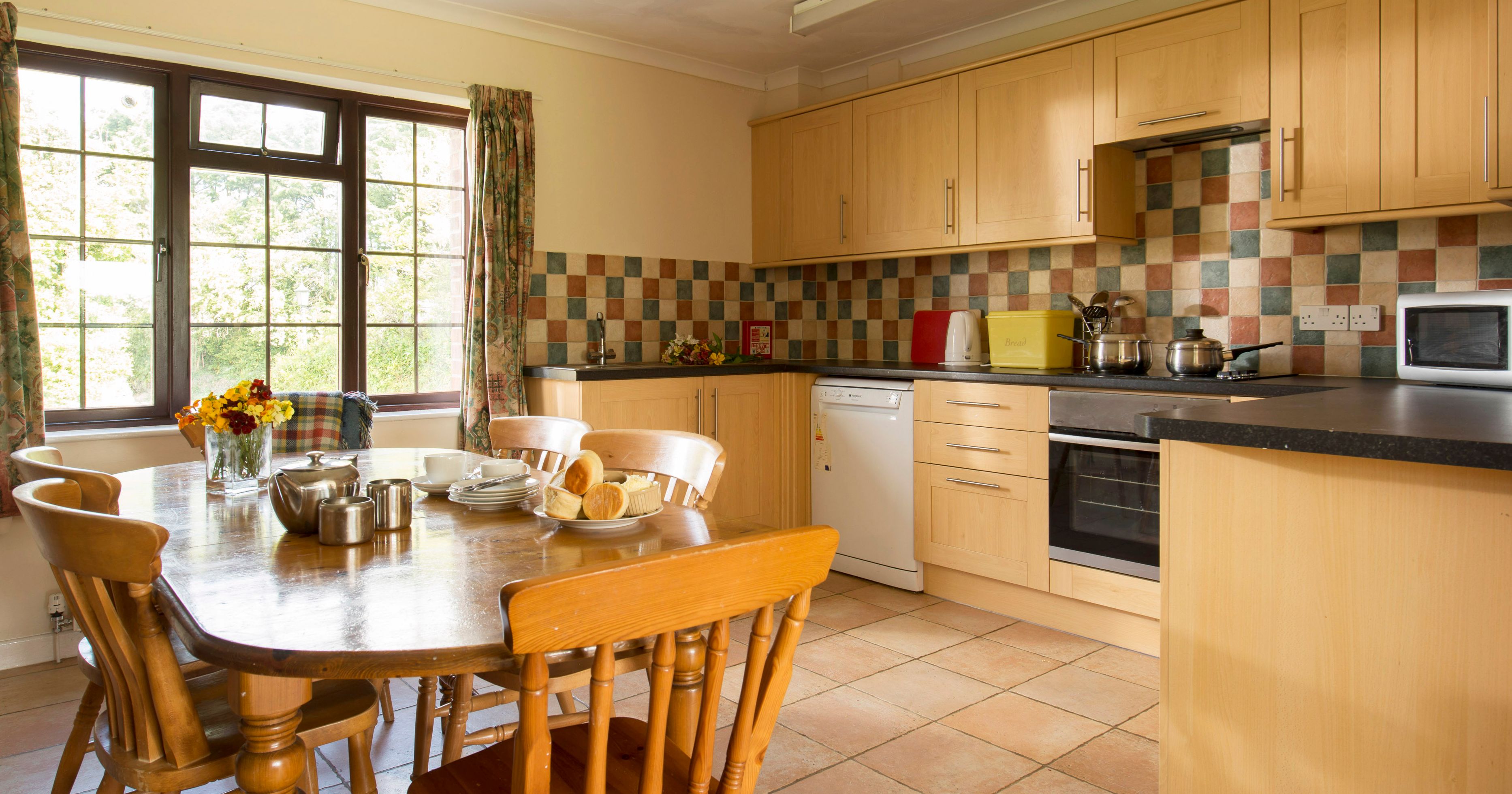 Cozy kitchen with wooden cabinets, a tiled backsplash, and a dining table set for tea with plates of pastries and a teapot. Large window provides natural light and view of greenery outside.