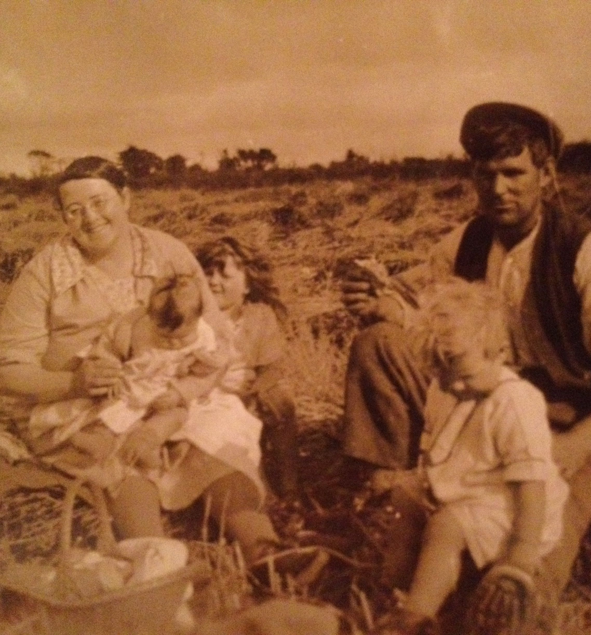 A sepia-toned vintage photograph of a family sitting in a field during harvest time. Two adults and three children are present, with a basket in the foreground.