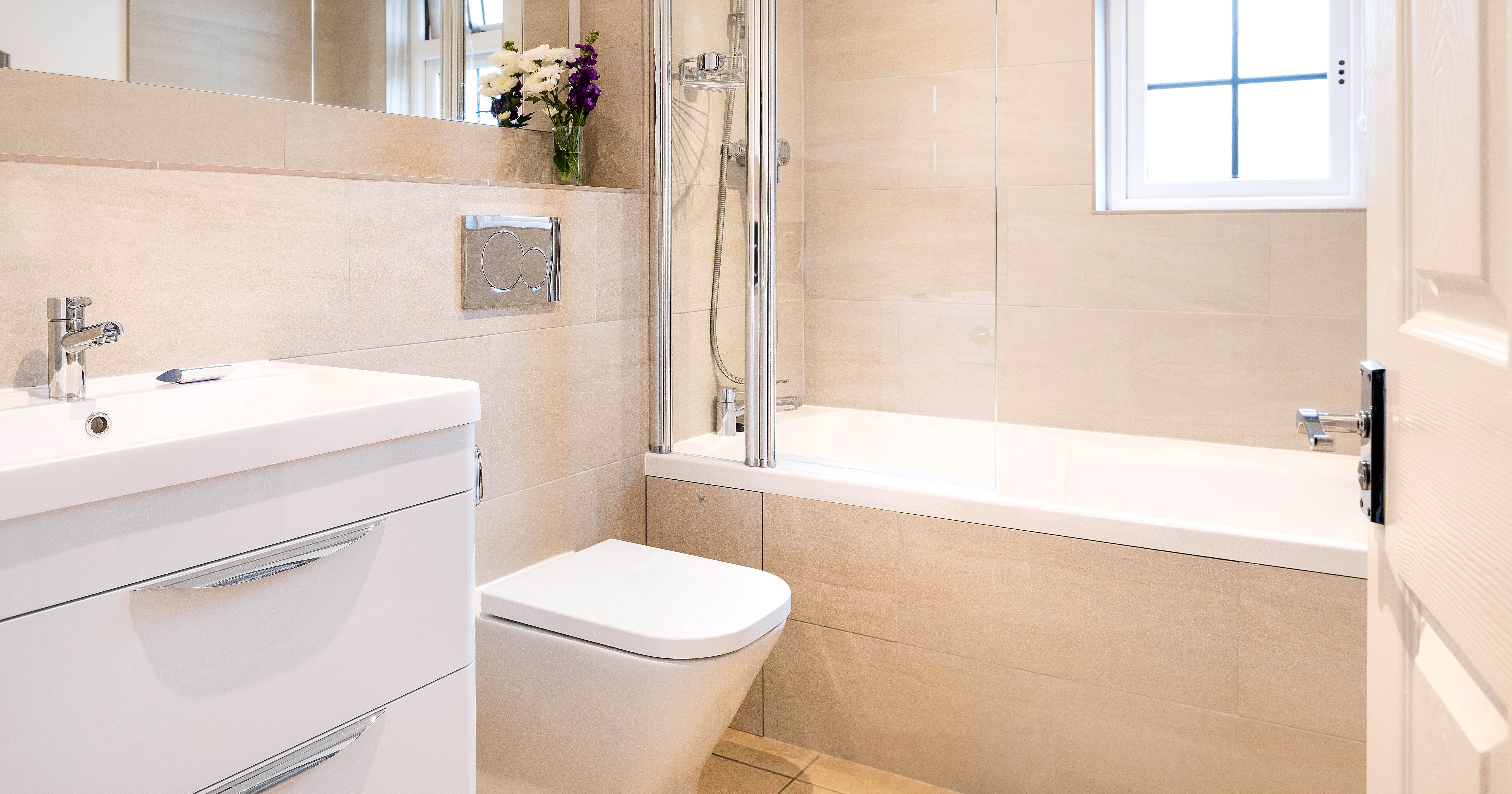Modern bathroom with beige tiles, a white sink with drawers, wall-mounted toilet, bathtub with glass shower screen, wall mirror, and a window.