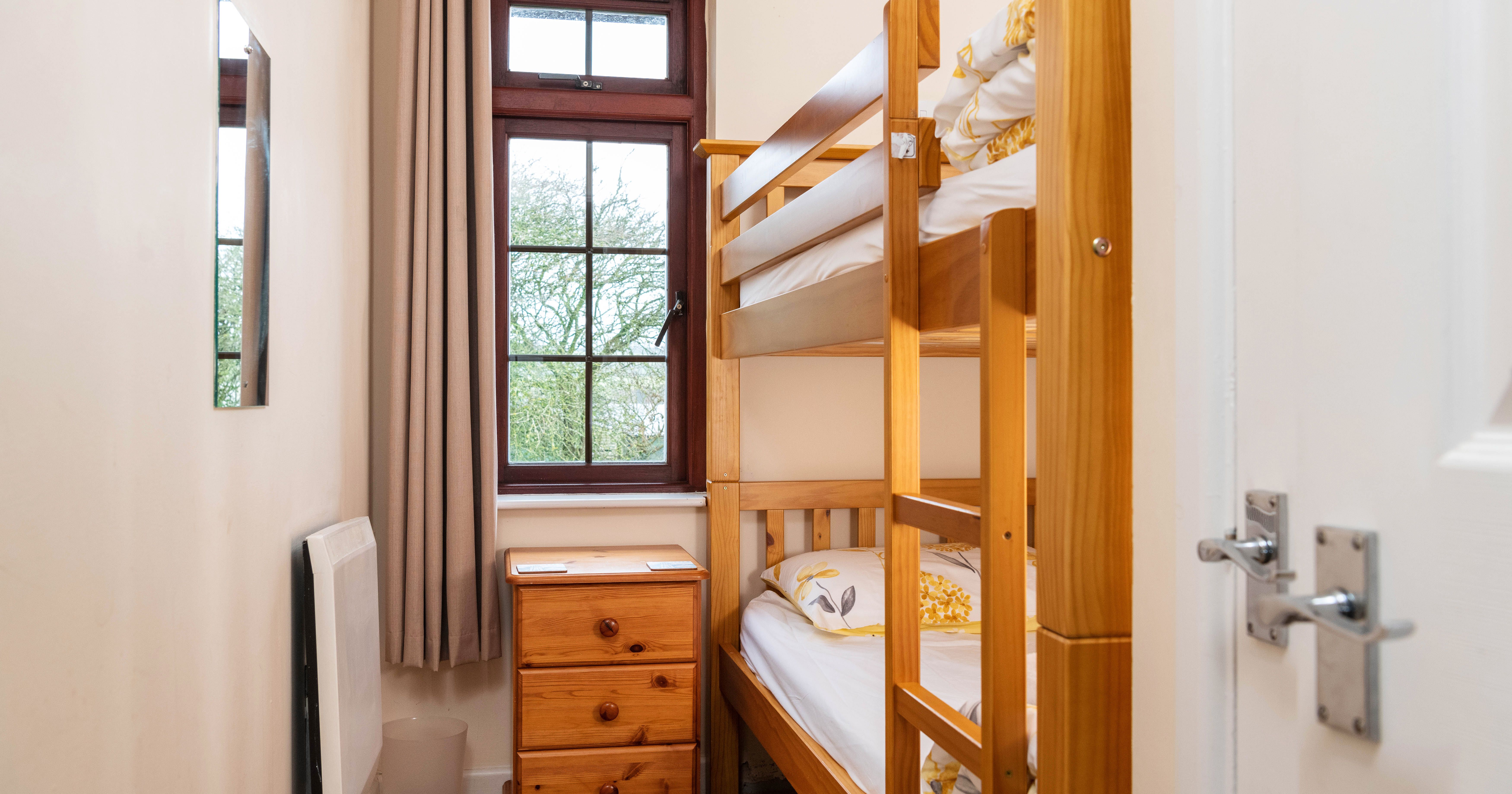 Small bedroom with wooden bunk beds, a wooden nightstand, a window with beige curtains, a wall mirror, and a white door.