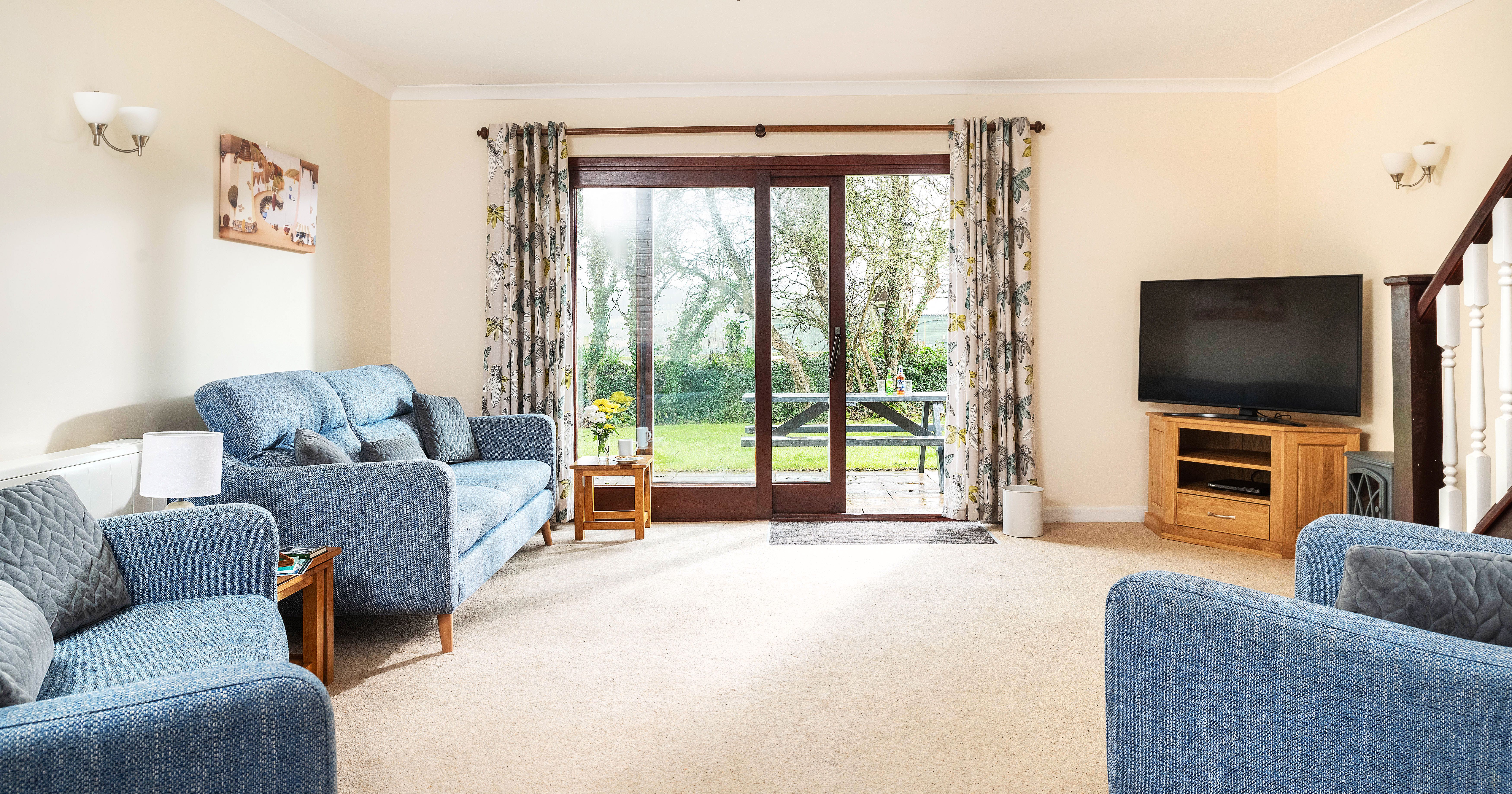 Bright and cozy living room with blue sofas, a large flat-screen TV on a wooden stand, beige carpet, floral curtains, and sliding glass doors leading to a green backyard with a picnic table.