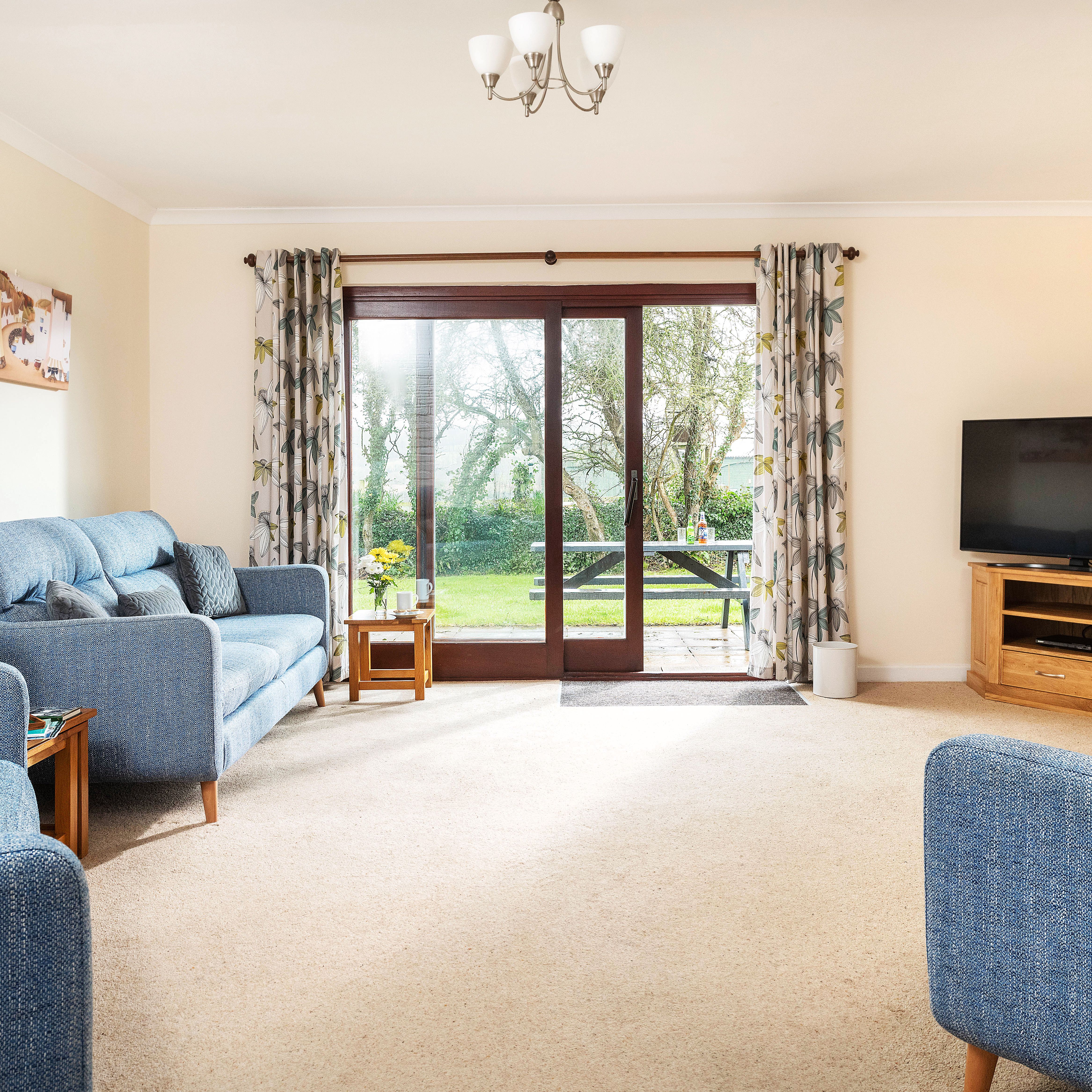 Bright and cozy living room with blue sofas, a large flat-screen TV on a wooden stand, beige carpet, floral curtains, and sliding glass doors leading to a green backyard with a picnic table.