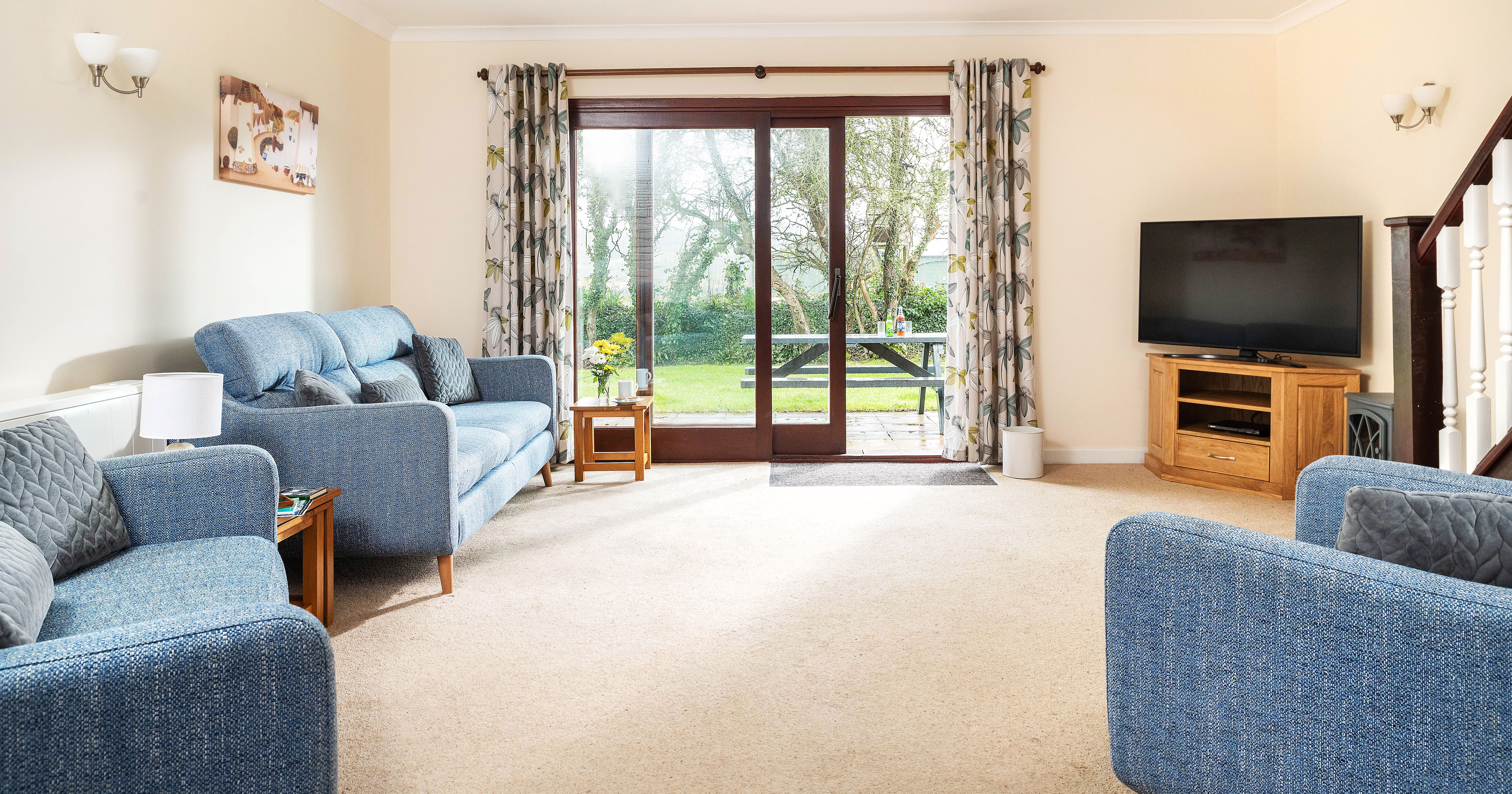 Bright and cozy living room with blue sofas, a large flat-screen TV on a wooden stand, beige carpet, floral curtains, and sliding glass doors leading to a green backyard with a picnic table.