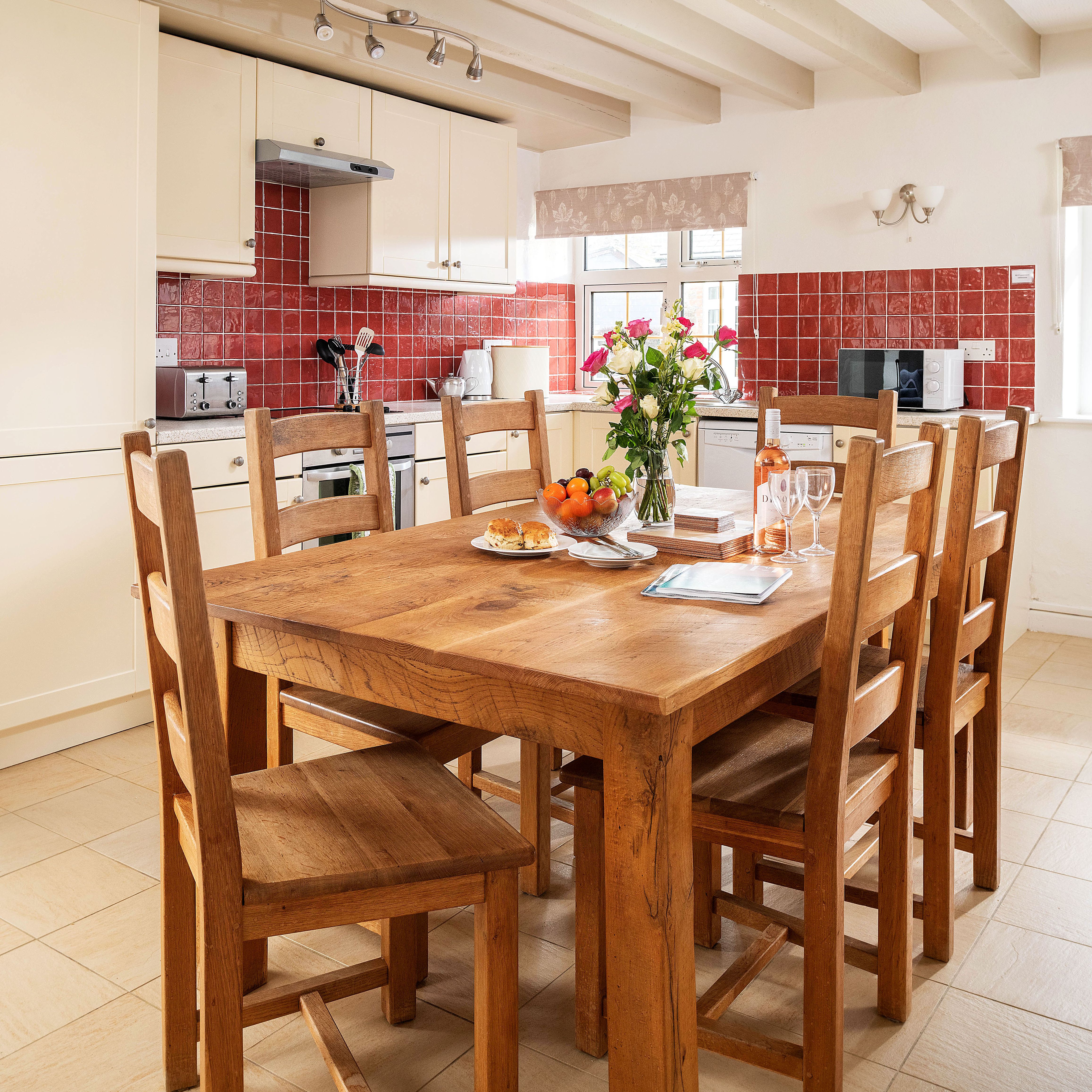 A bright and cozy kitchen with cream cabinets, red tiled backsplash, and wooden dining table set for a meal with flowers, fruit, and wine. Two jackets hang on hooks on the wall.