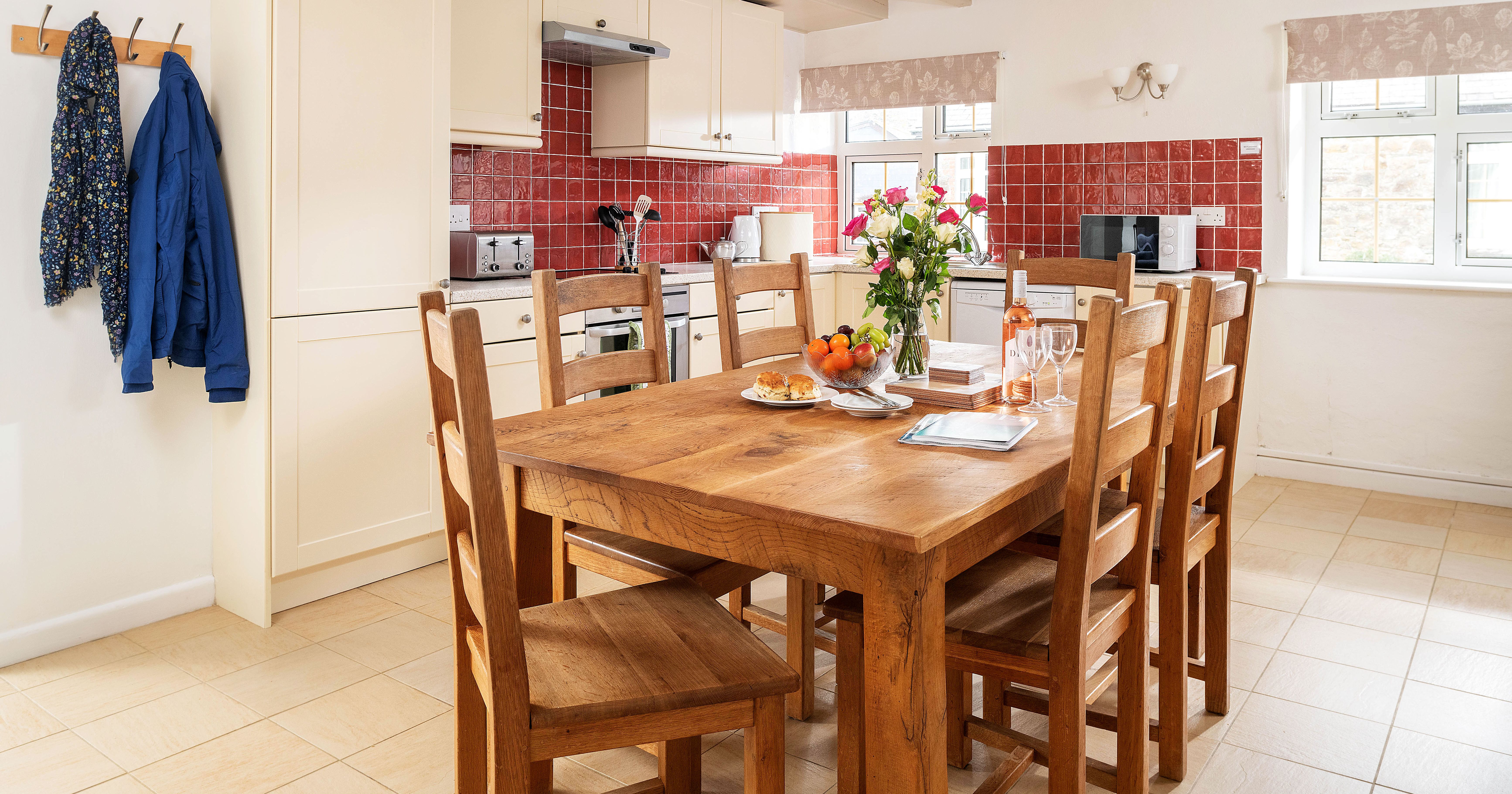 A bright and cozy kitchen with cream cabinets, red tiled backsplash, and wooden dining table set for a meal with flowers, fruit, and wine. Two jackets hang on hooks on the wall.