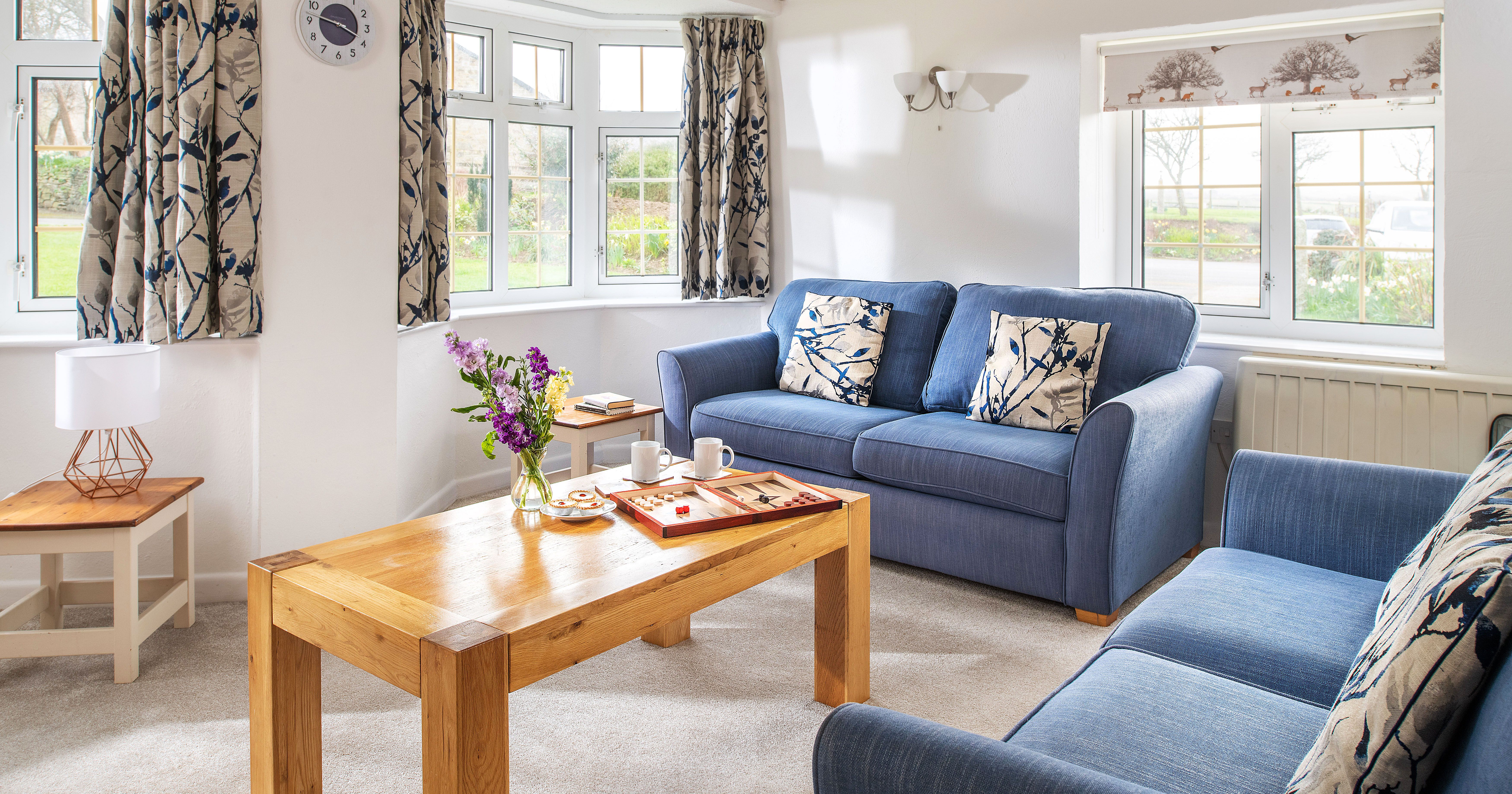 Cozy living room with two blue sofas, floral cushions, wooden coffee table with board game and snacks, large windows with patterned curtains, and a table lamp on a side table.
