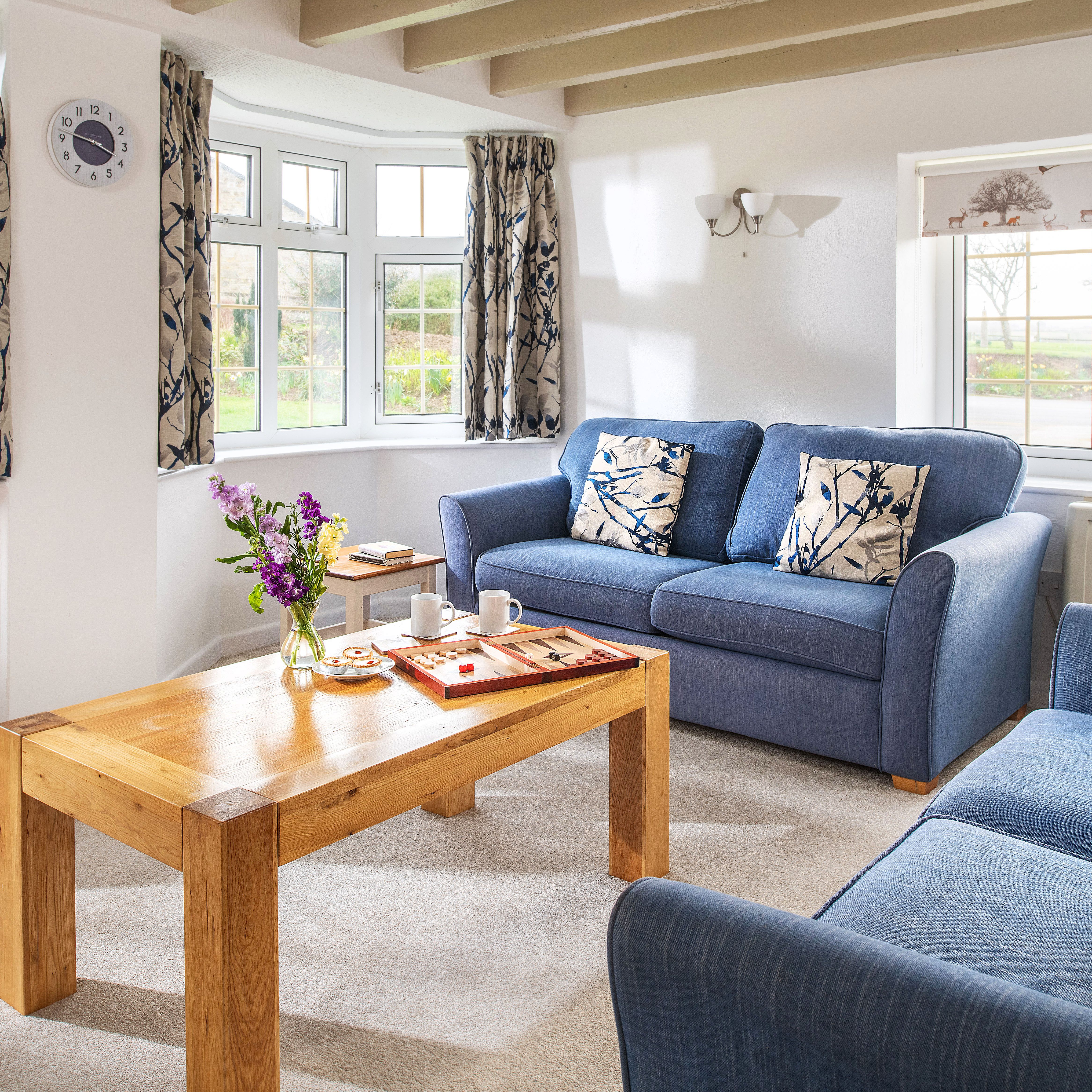 Cozy living room with two blue sofas, floral cushions, wooden coffee table with board game and snacks, large windows with patterned curtains, and a table lamp on a side table.