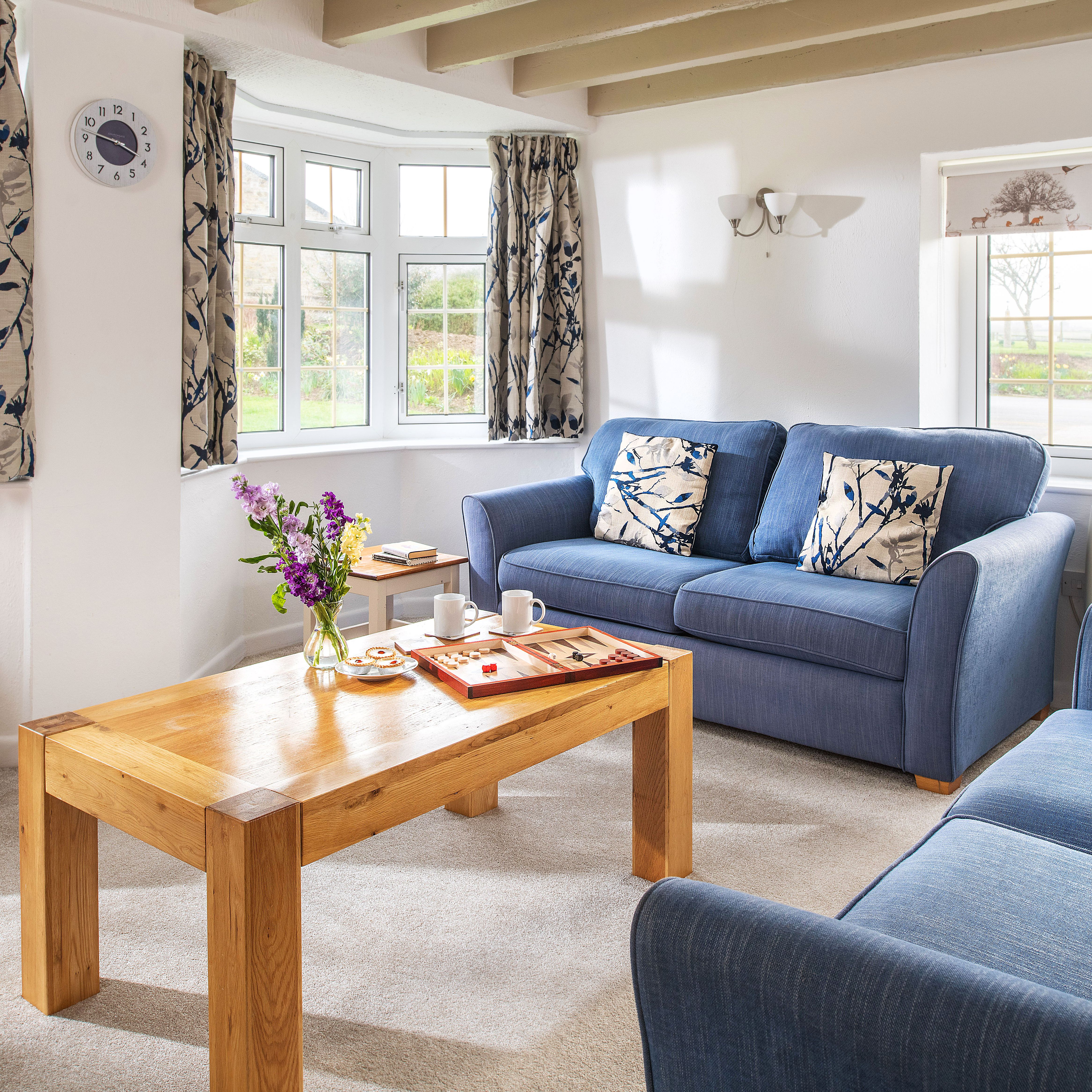 Cozy living room with two blue sofas, floral cushions, wooden coffee table with board game and snacks, large windows with patterned curtains, and a table lamp on a side table.