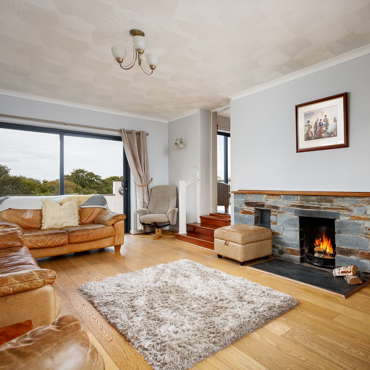 A cozy living room with wooden floors, tan leather sofas, a fluffy rug, a stone fireplace with a burning fire, a large window with curtains, and a framed picture above the fireplace.