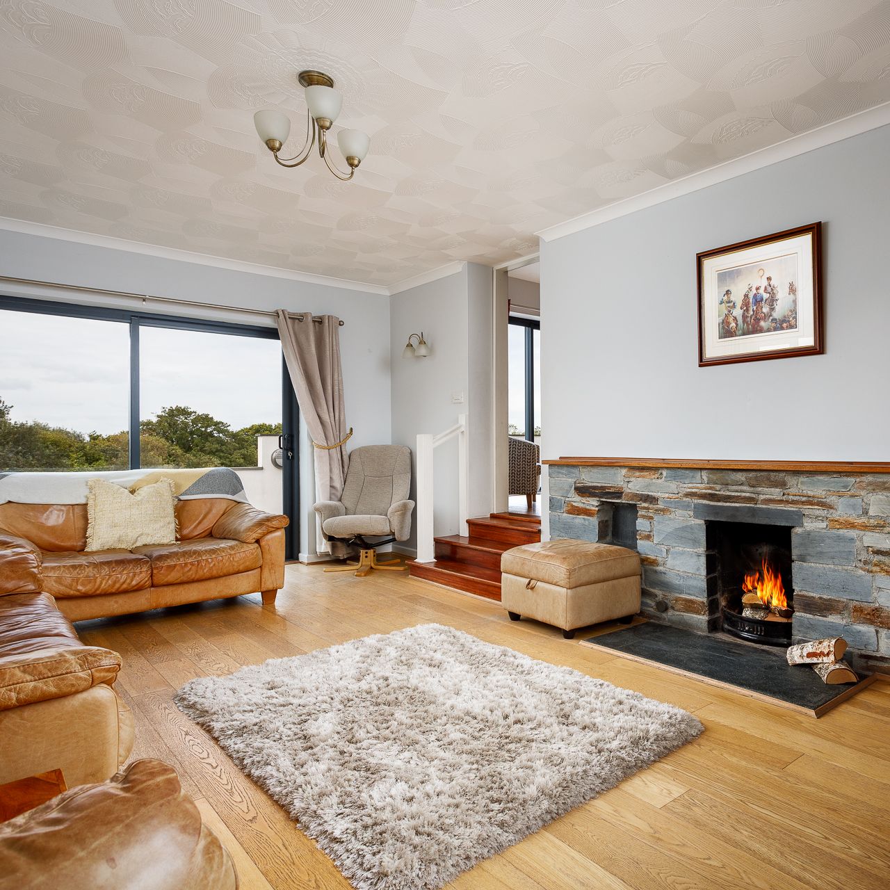 A cozy living room with wooden floors, tan leather sofas, a fluffy rug, a stone fireplace with a burning fire, a large window with curtains, and a framed picture above the fireplace.
