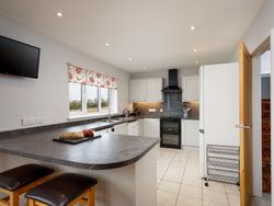 Modern kitchen with white cabinets, black countertops, a central island with two stools, wall-mounted TV, window with patterned blind, and stainless steel appliances.