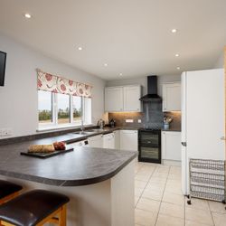 Modern kitchen with white cabinets, black countertops, a central island with two stools, wall-mounted TV, window with patterned blind, and stainless steel appliances.