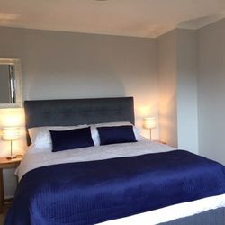 Modern bedroom with a grey upholstered bed, navy blue bedding, two matching bedside tables with lamps, a wall mirror on the left, and floral wall art on the right.