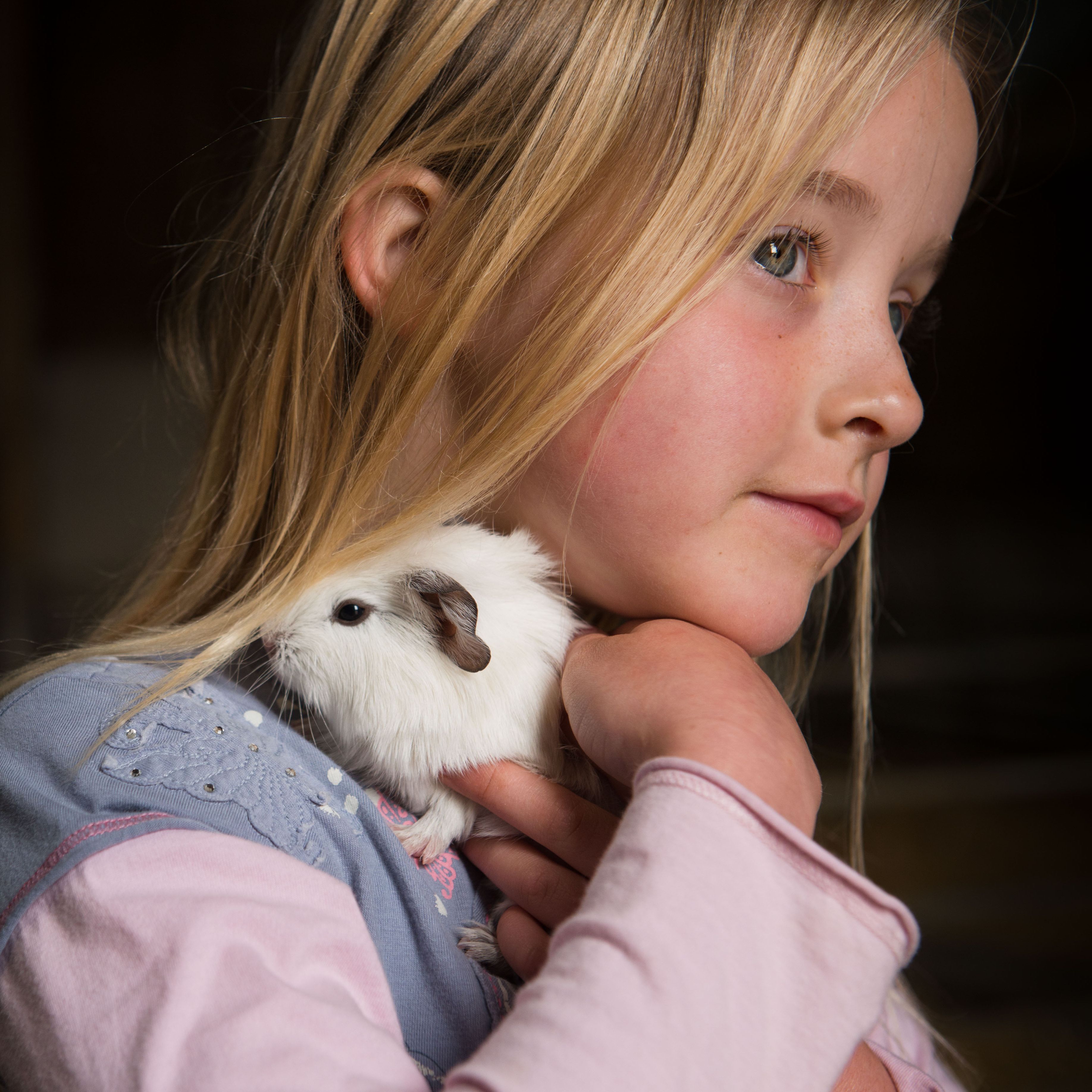 A young girl with blonde hair holding a white guinea pig close to her neck, with the guinea pig partly hidden under her hair.