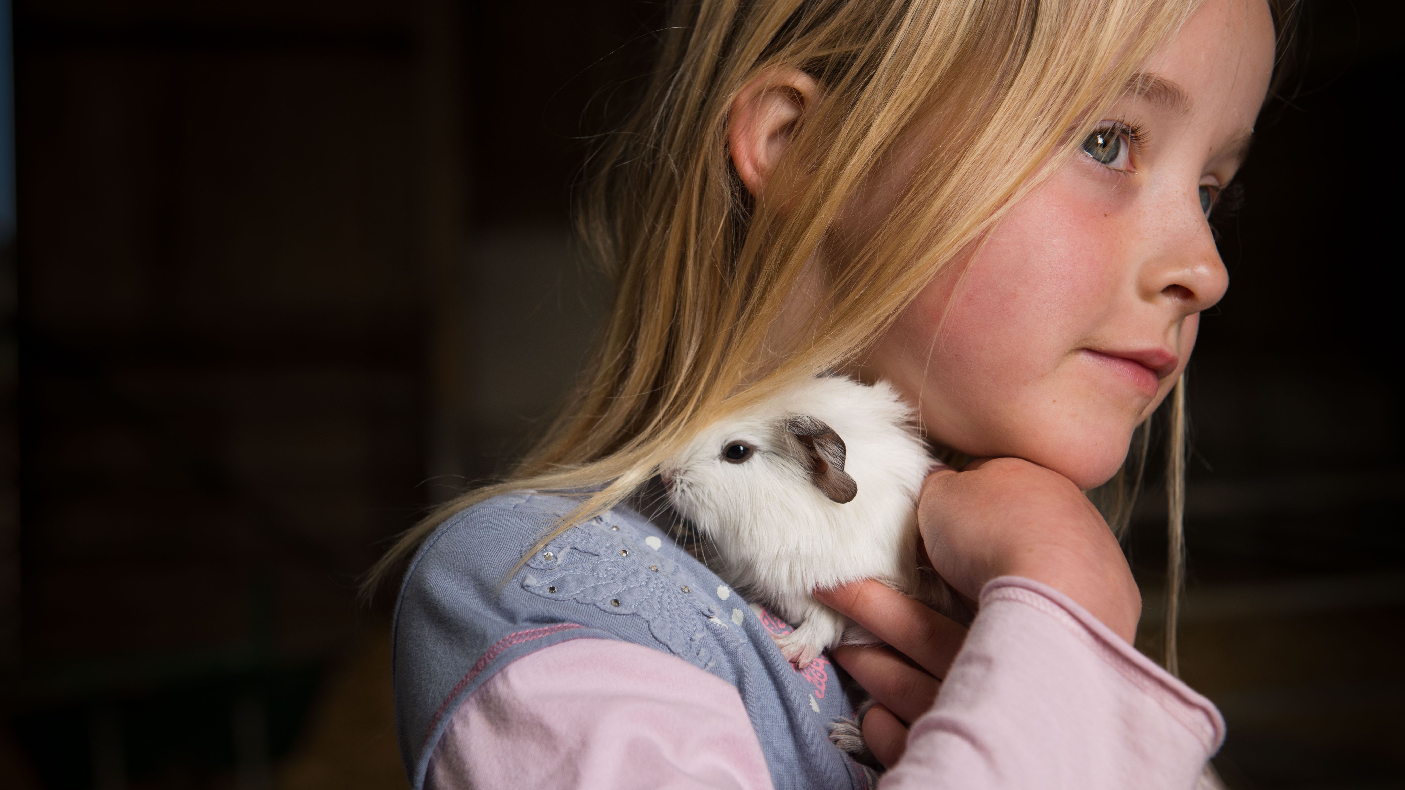 A young girl with blonde hair holding a white guinea pig close to her neck, with the guinea pig partly hidden under her hair.