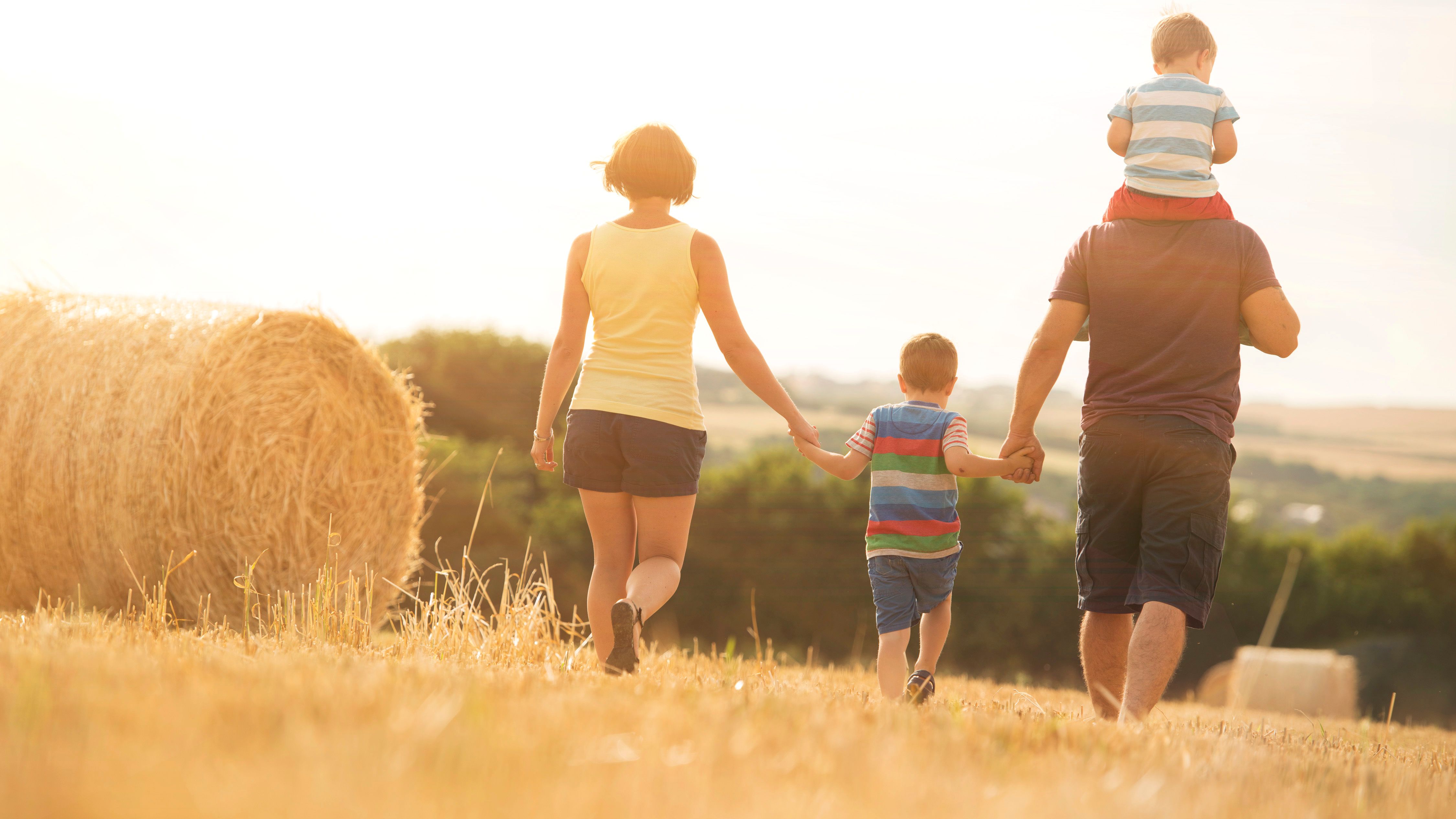 Family of four walking in a sunny field with hay bales, holding hands, with one child riding on the father's shoulders.