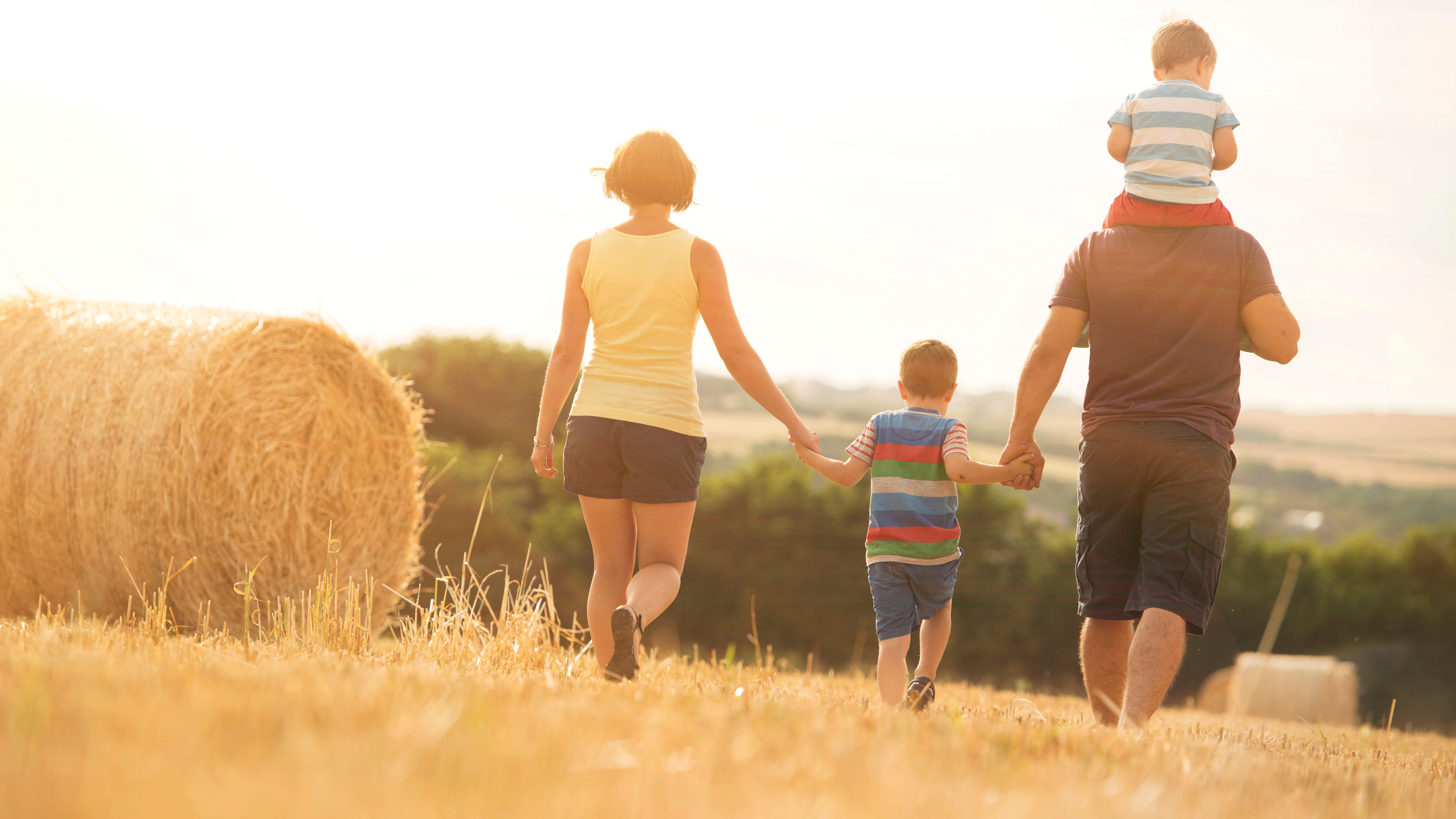 Family of four walking in a sunny field with hay bales, holding hands, with one child riding on the father's shoulders.
