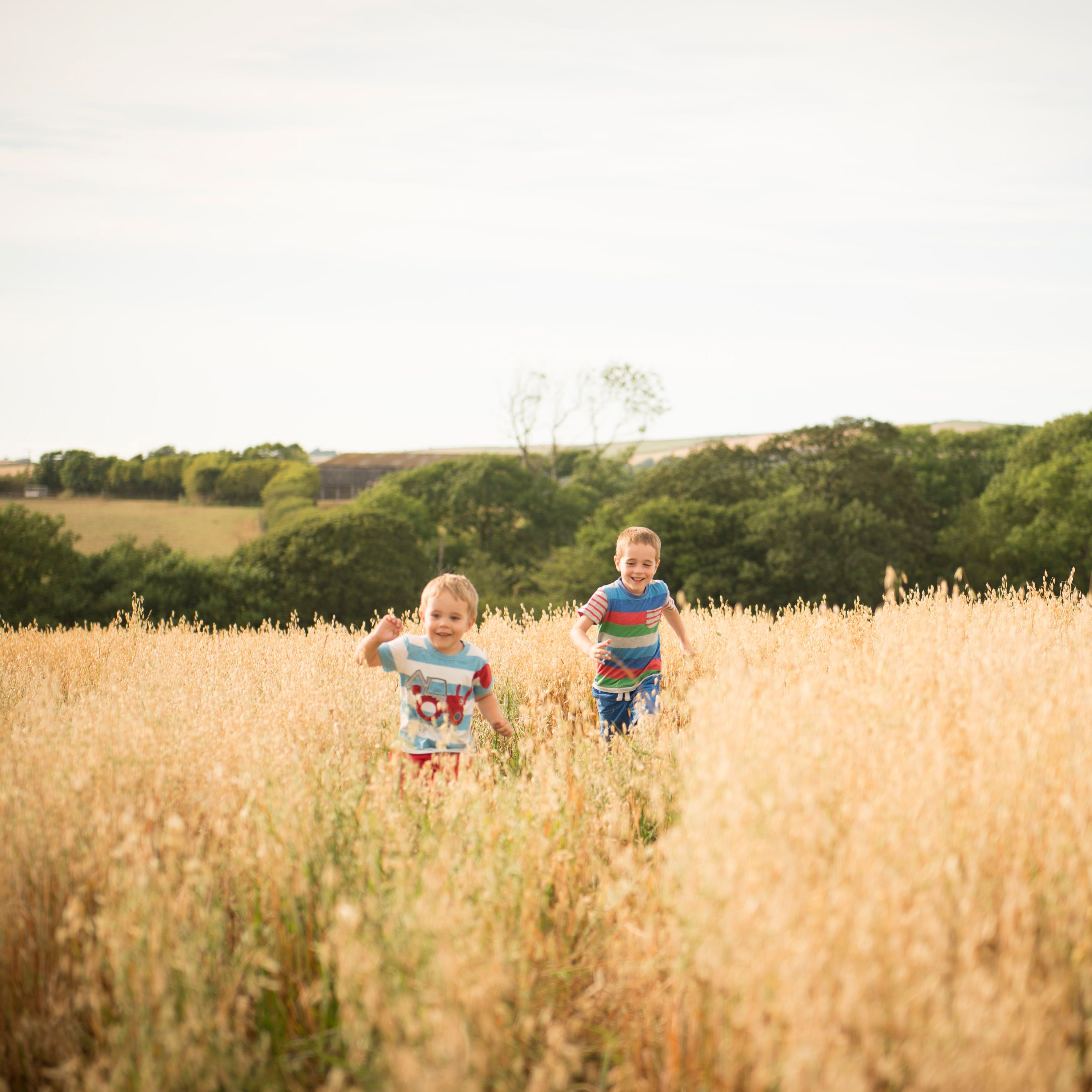 Two children running and smiling in a tall grassy field with trees and hills in the background.