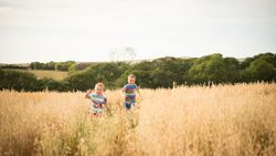 Two children running and smiling in a tall grassy field with trees and hills in the background.