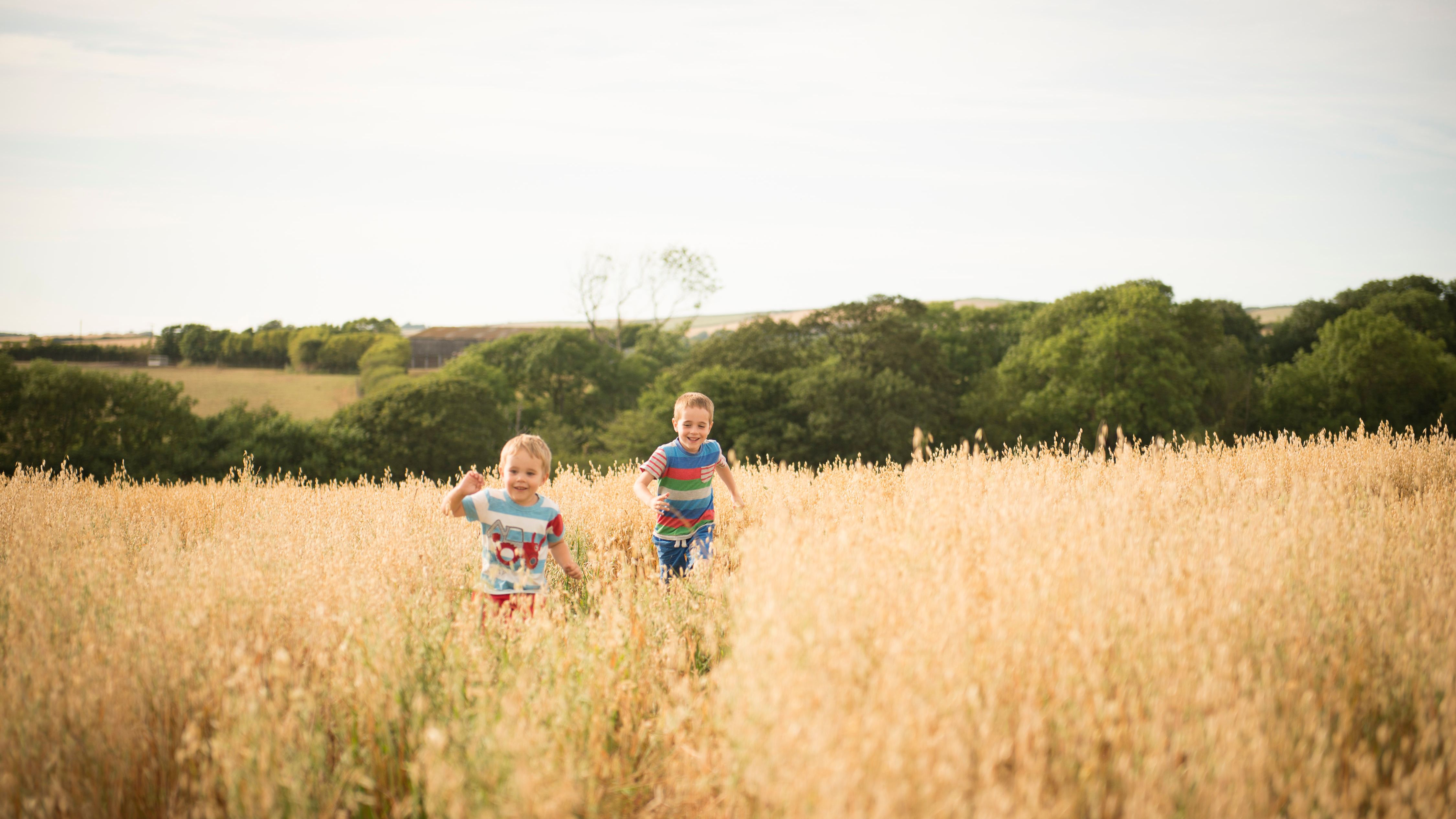 Two children running and smiling in a tall grassy field with trees and hills in the background.