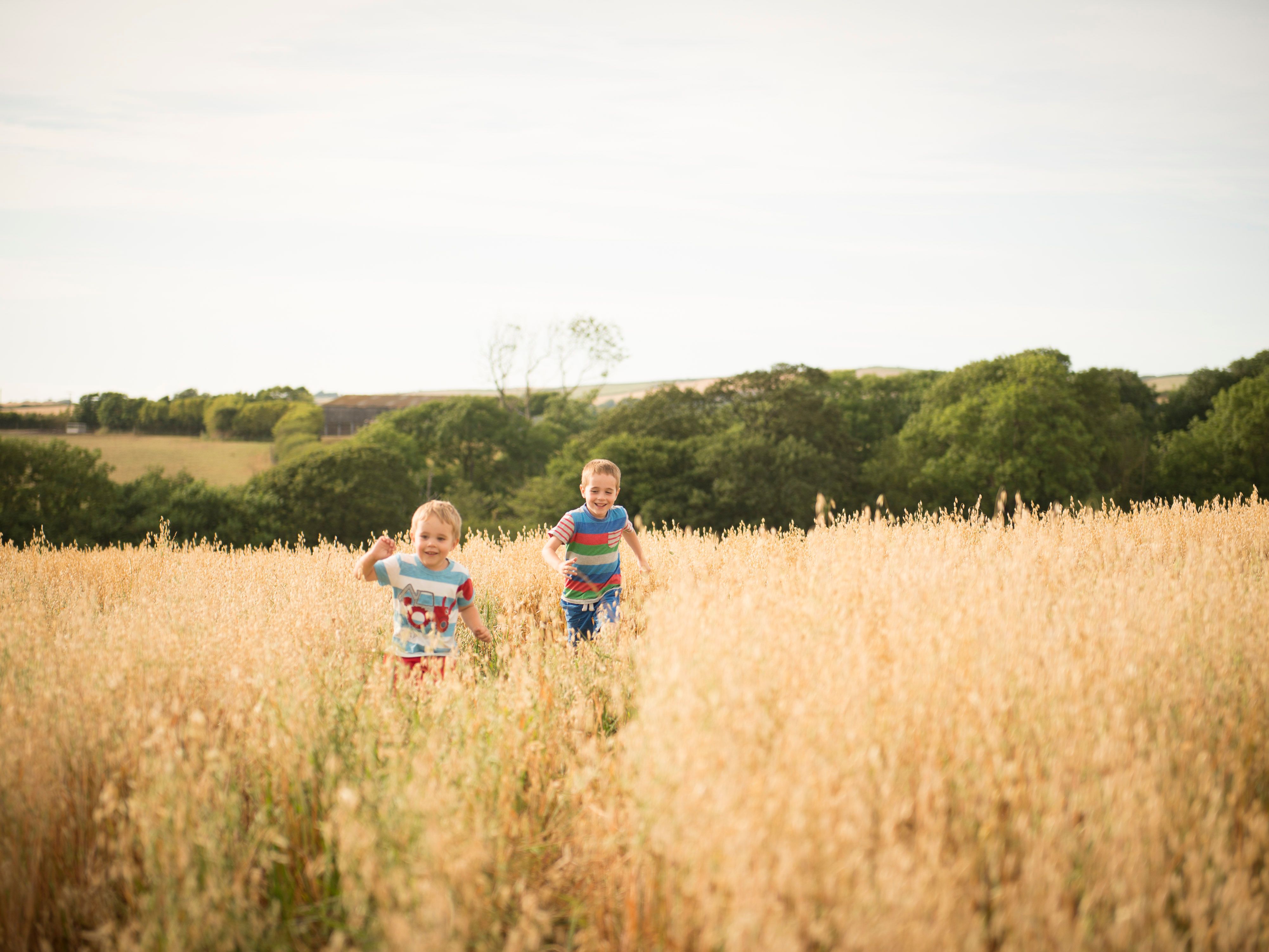 Two children running and smiling in a tall grassy field with trees and hills in the background.