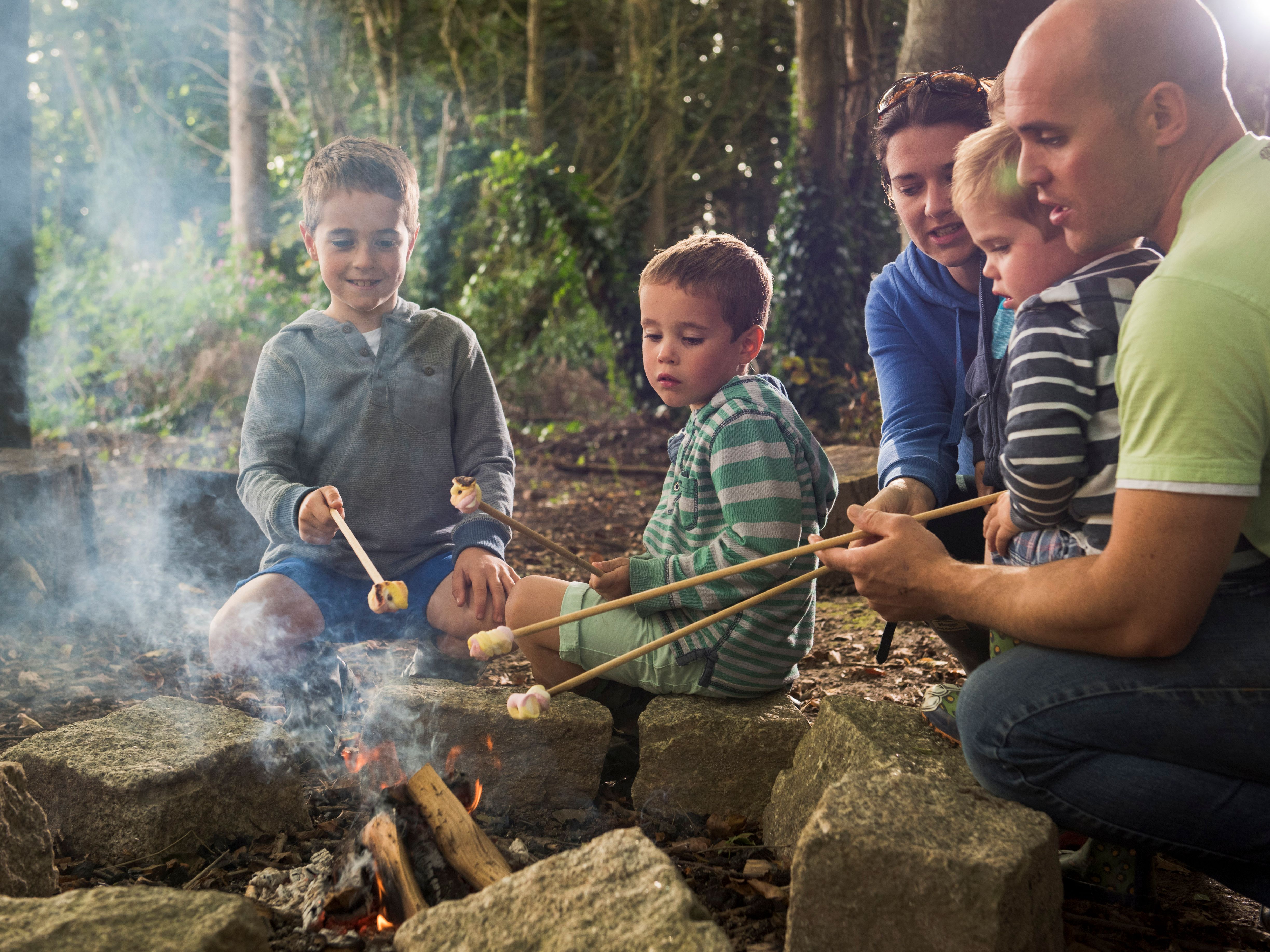 A family sitting around a campfire in the woods, roasting marshmallows on sticks. Two adults and three young children are enjoying the outdoor activity together.