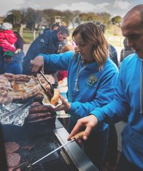 People wearing blue hoodies are grilling and serving food at an outdoor barbecue event, with a group of people and children waiting in the background.