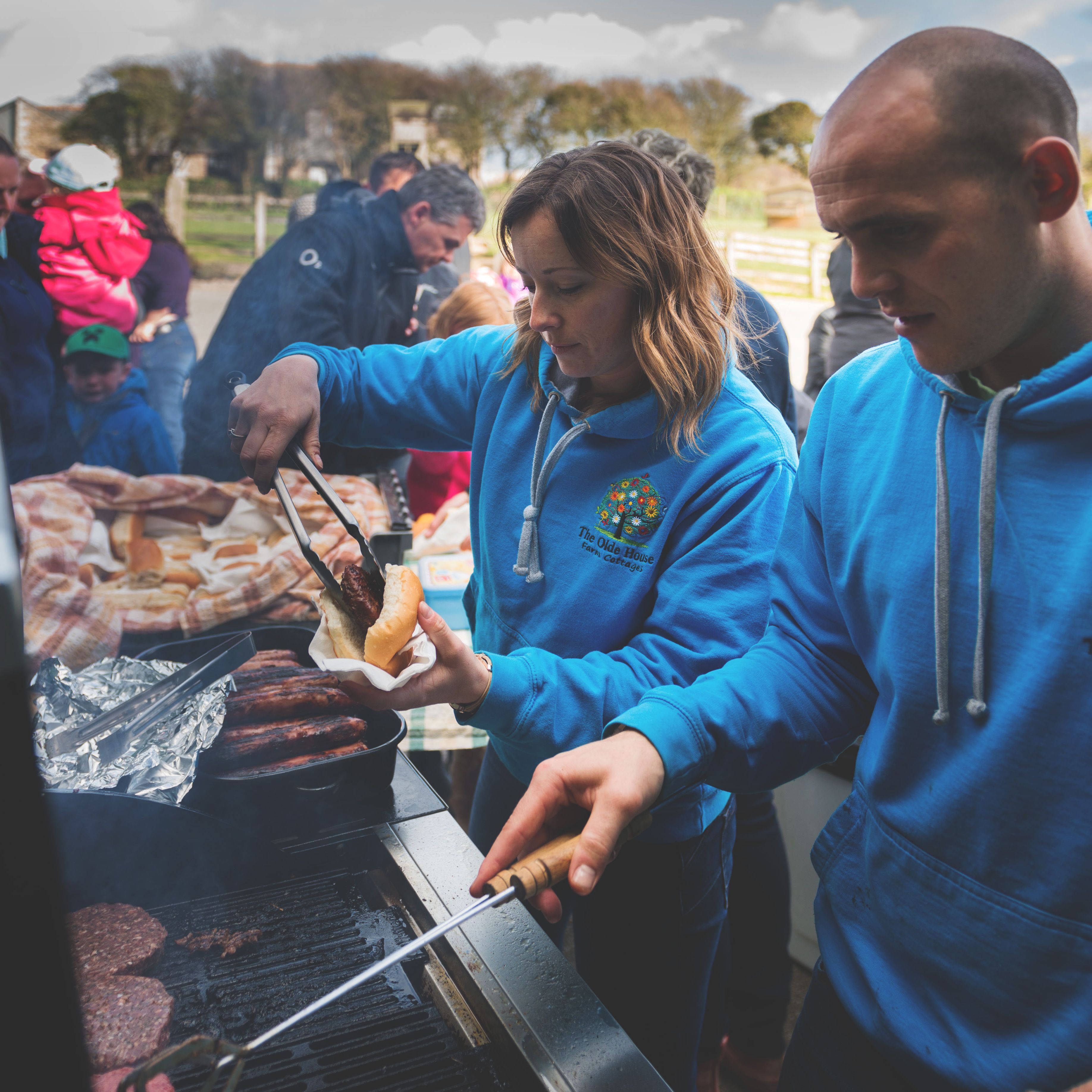 People wearing blue hoodies are grilling and serving food at an outdoor barbecue event, with a group of people and children waiting in the background.